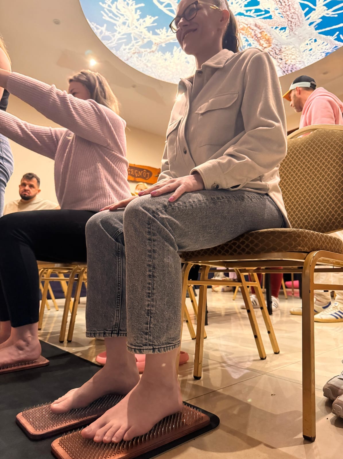 Uczestnicy siedzą na krzesłach podczas sesji relaksacyjnej z deskami z gwoździami, w tle widoczne osoby i sufit z motywem nieba.