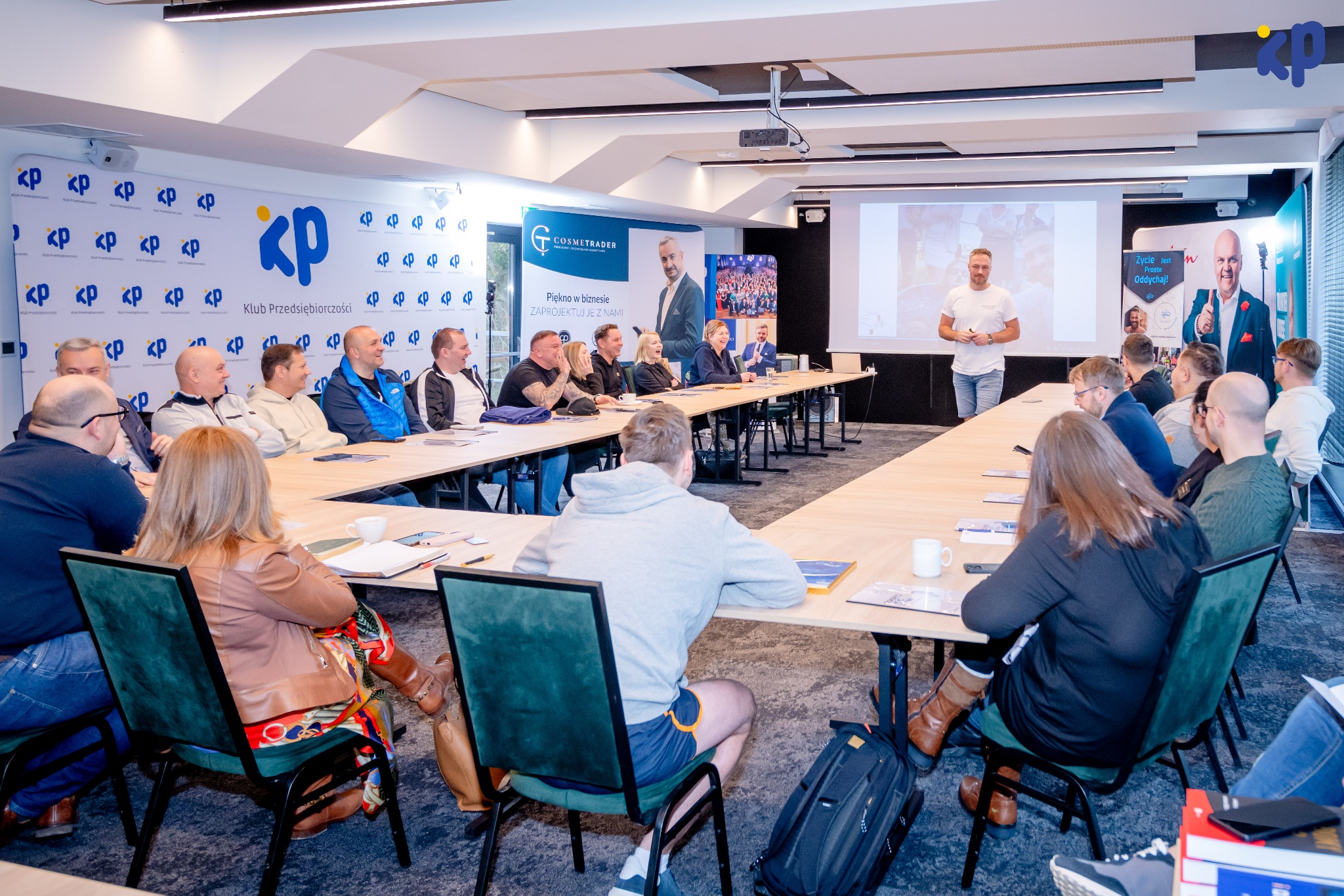 Prelegent w białej koszulce prowadzi prezentację dla grupy osób siedzących przy długim stole w sali konferencyjnej z logo Klubu Przedsiębiorczości w tle.