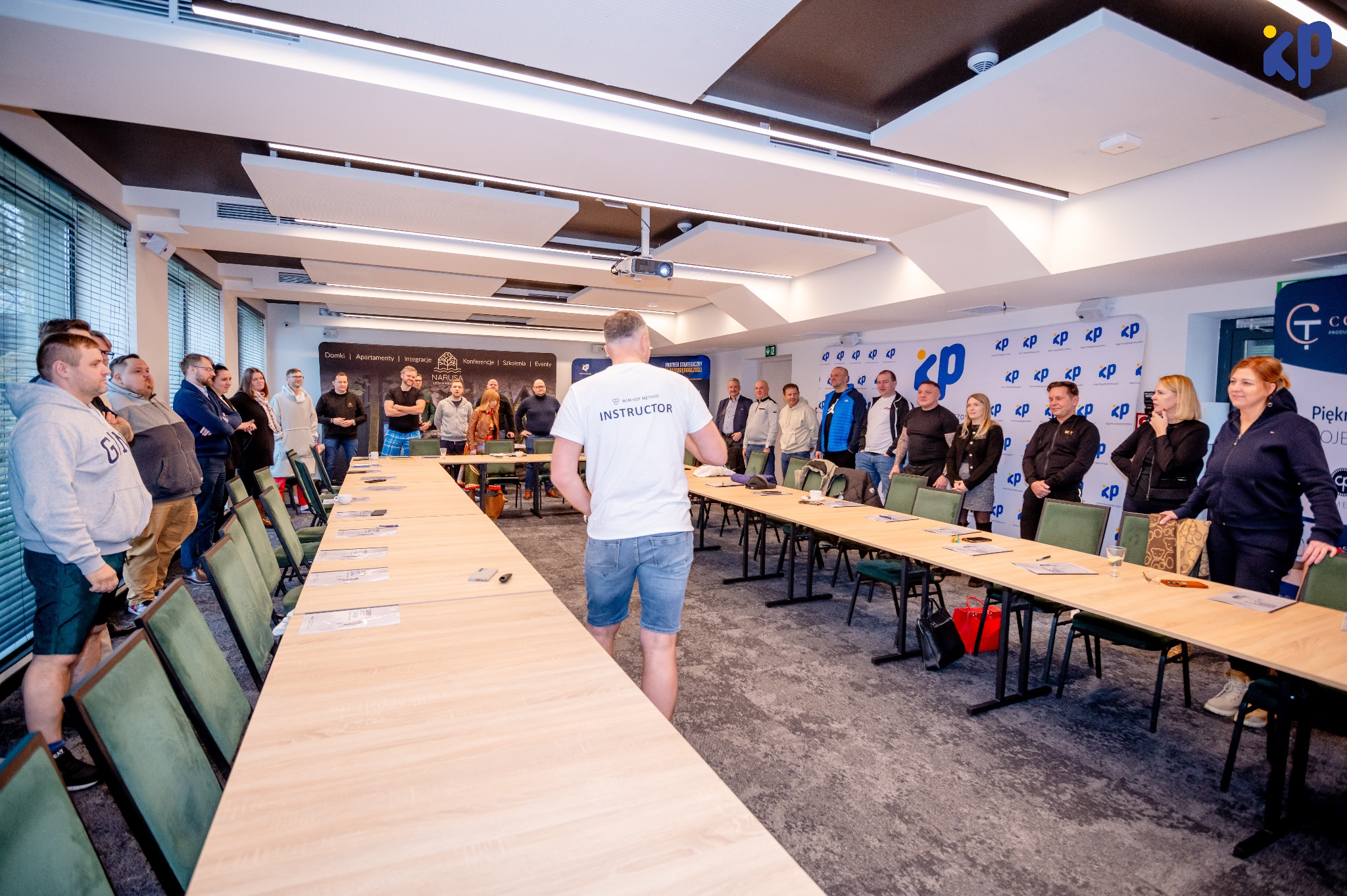Sala konferencyjna: instruktor prowadzi szkolenie dla grupy osób. Widoczne stoły, krzesła i uczestnicy. Na ścianie logo firmy. Instruktor w koszulce z napisem INSTRUCTOR.