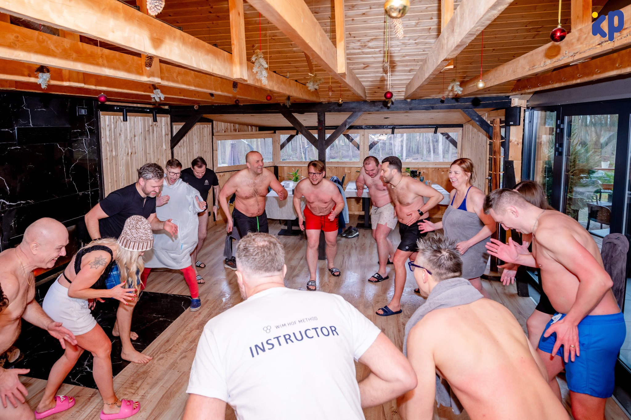 Grupa osób, częściowo rozebranych, uczestniczy w zajęciach prowadzonych przez instruktora Wim Hof Method w drewnianym wnętrzu z dekoracjami świątecznymi.