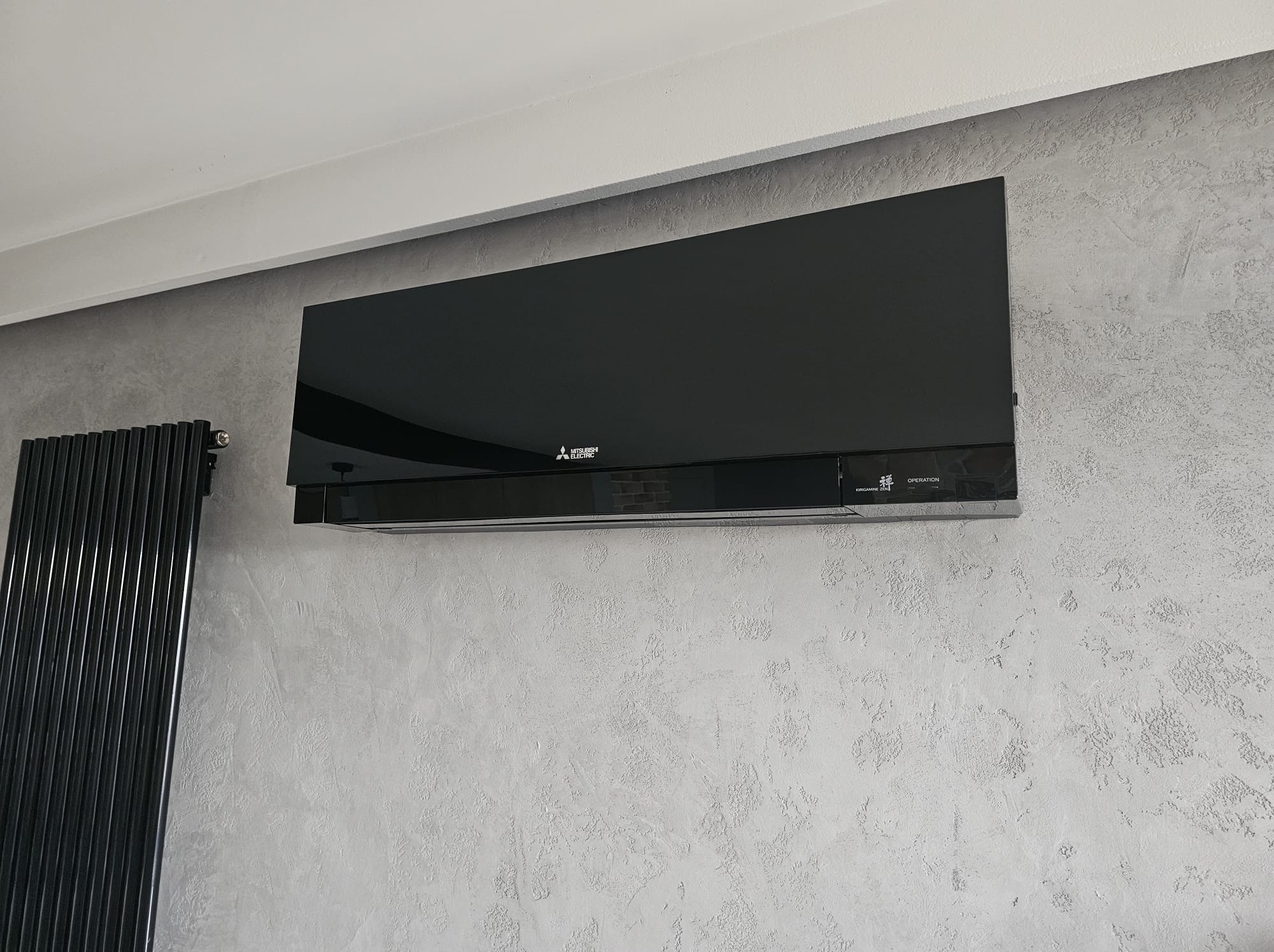 Czarny, nowoczesny klimatyzator Mitsubishi Electric na szarej ścianie o fakturze betonu, obok grzejnik żeberkowy. Minimalistyczny design, eleganckie wykończenie.