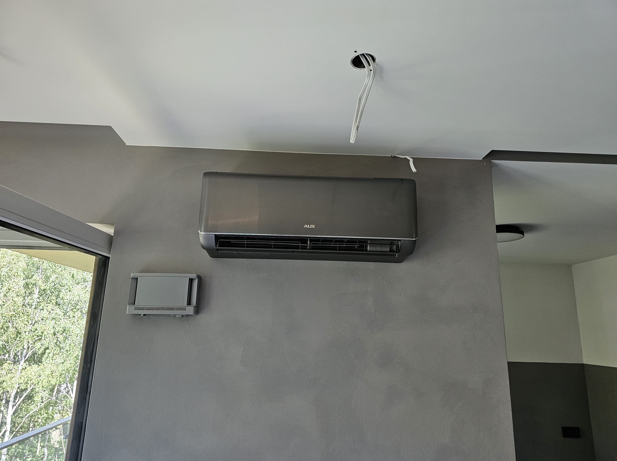 Szary klimatyzator AUX na ścianie o fakturze betonu, z widocznymi przewodami wychodzącymi z sufitu. Obok okno z widokiem na zieleń. Minimalistyczny design wnętrza.