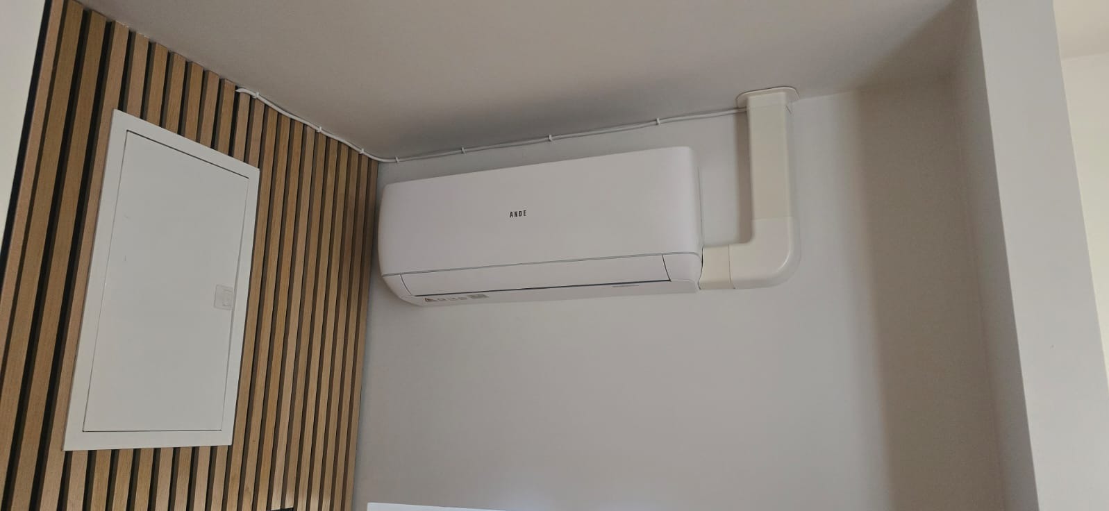 Biały klimatyzator marki ANDE zamontowany na ścianie, z widocznymi przewodami i elementami instalacji. Obok skrzynka elektryczna na tle drewnianej ściany.