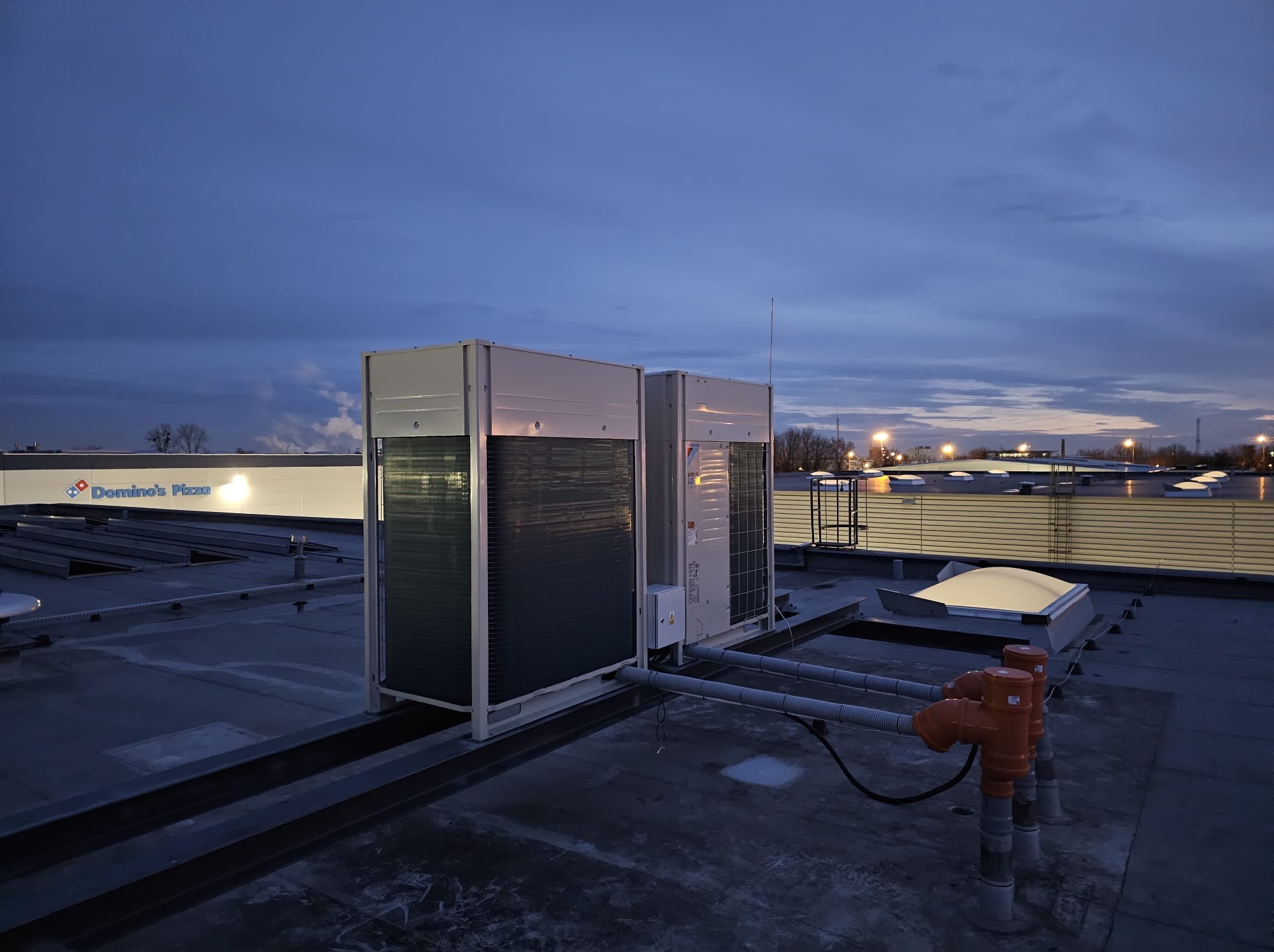 Dwa klimatyzatory na dachu budynku komercyjnego, widoczne rury i elementy instalacji. W tle budynek Domino's Pizza oświetlony wieczornym światłem.