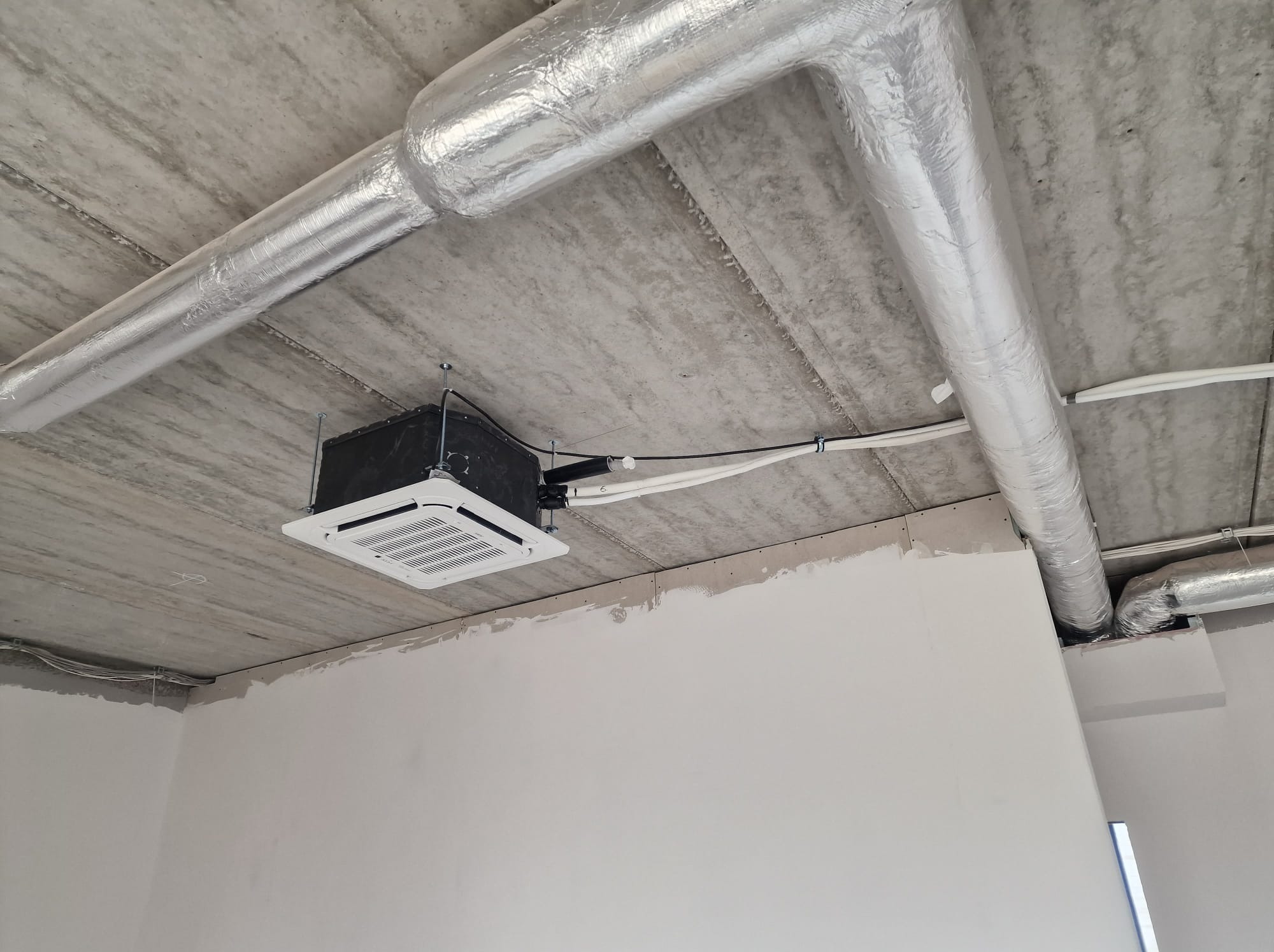 Kasetonowa jednostka klimatyzacji zamontowana pod betonowym stropem, widoczne rury w srebrnej izolacji i przewody. Ściany w trakcie wykańczania.