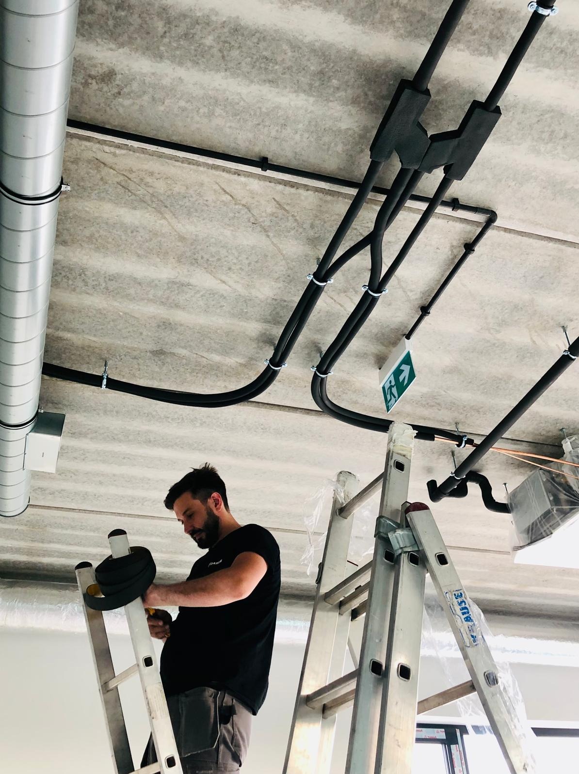 Instalator na drabinie montuje rury wentylacyjne pod betonowym sufitem. Widoczne elementy instalacji, rury w izolacji, kanał wentylacyjny i znak ewakuacyjny.