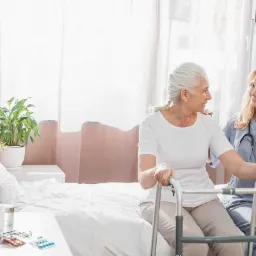 Uśmiechnięta starsza kobieta z siwymi włosami siedzi na łóżku, trzymając się chodzika. Obok niej stoi pielęgniarka w niebieskim uniformie i ze stetoskopem na szyi, trzymając ją za rękę. Na stoliku...