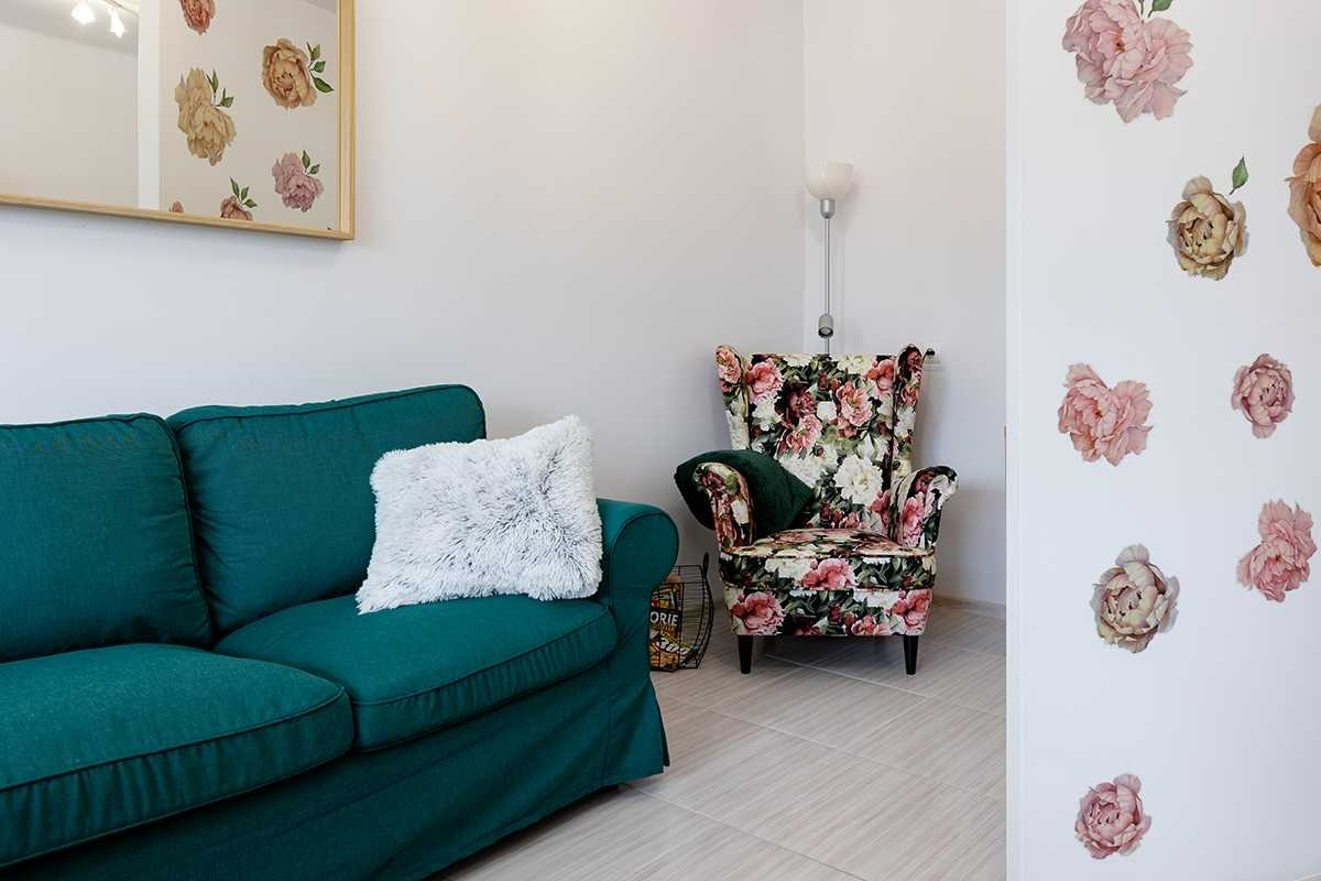 Aranżacja wnętrza: szmaragdowa sofa z puszystą poduszką, fotel w kwiaty i naklejki kwiatowe na ścianie.