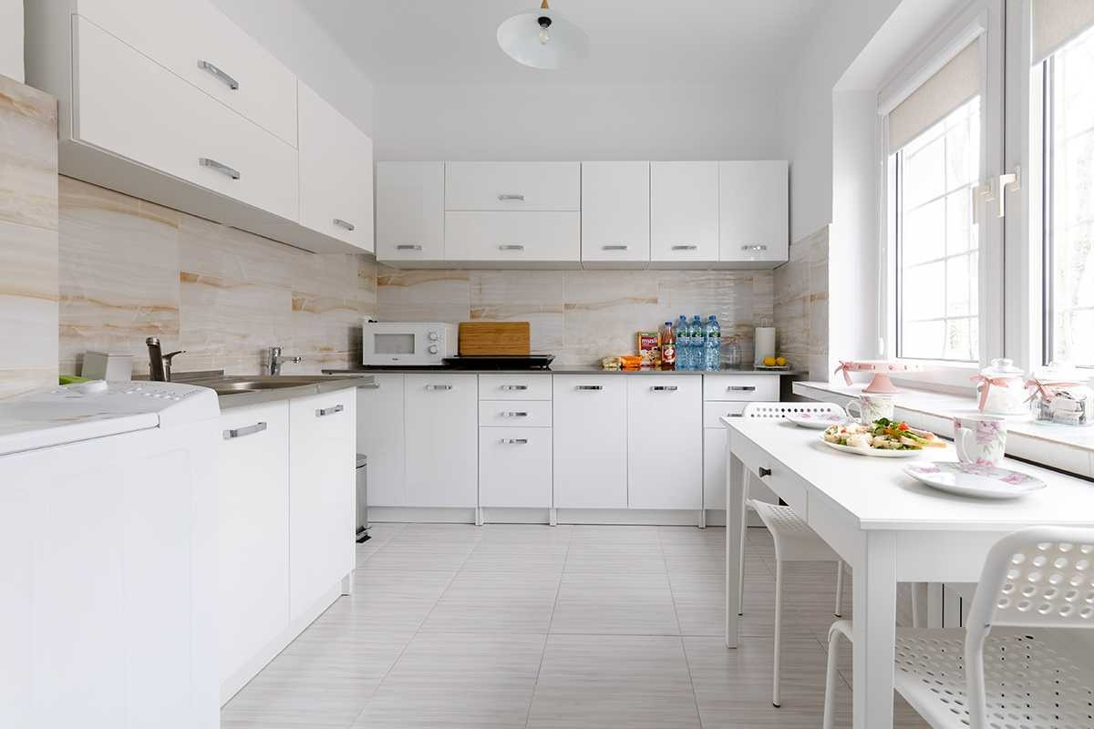 Jasna kuchnia z białymi meblami, marmurowym blatem i stołem z nakryciem, przy oknie dekoracje z różowymi wstążkami.