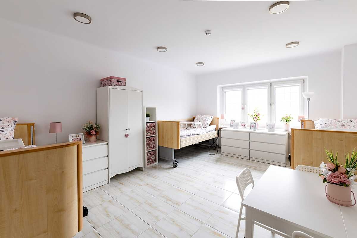 Jasny pokój z dwoma łóżkami rehabilitacyjnymi na kółkach, szafą, komodami i stolikiem z krzesłami, kwiaty w wazonach i ramki na zdjęciach na komodach, podłoga wyłożona płytkami imitującymi marmur.