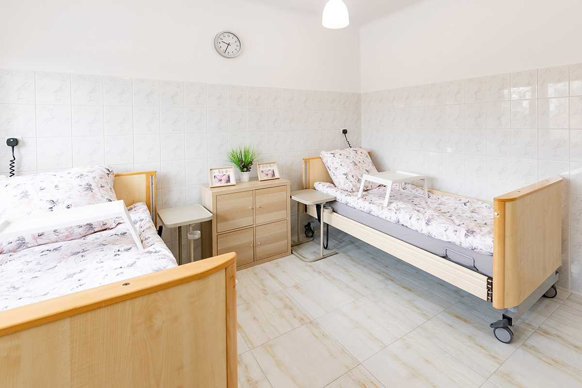 Dwupokojowy pokój w jasnych barwach z dwoma łóżkami szpitalnymi na kółkach, szafką nocną, stolikiem i dekoracjami w ramkach.