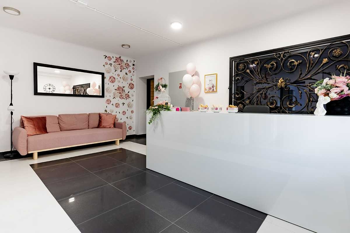 Jasne, przestronne wnętrze recepcji z białym biurkiem, różową sofą z aksamitnymi poduszkami, kwiatowymi dekoracjami na ścianie i ozdobnym metaloplastycznym panelem za biurkiem.