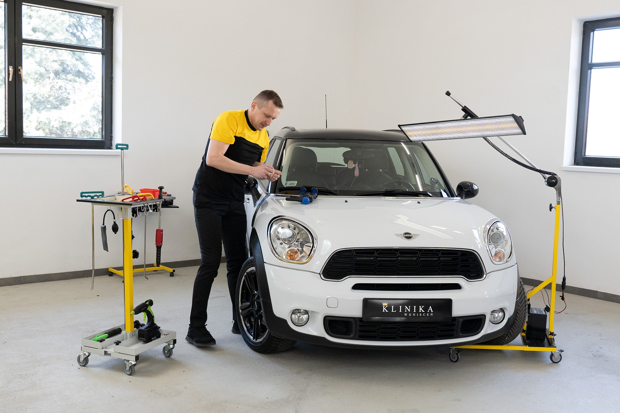 Pracownik w czarno-żółtej koszulce usuwa wgniecenie na białym Mini Cooperze, używając specjalistycznych narzędzi i lampy inspekcyjnej na mobilnym stojaku. W tle widoczny stół warsztatowy...