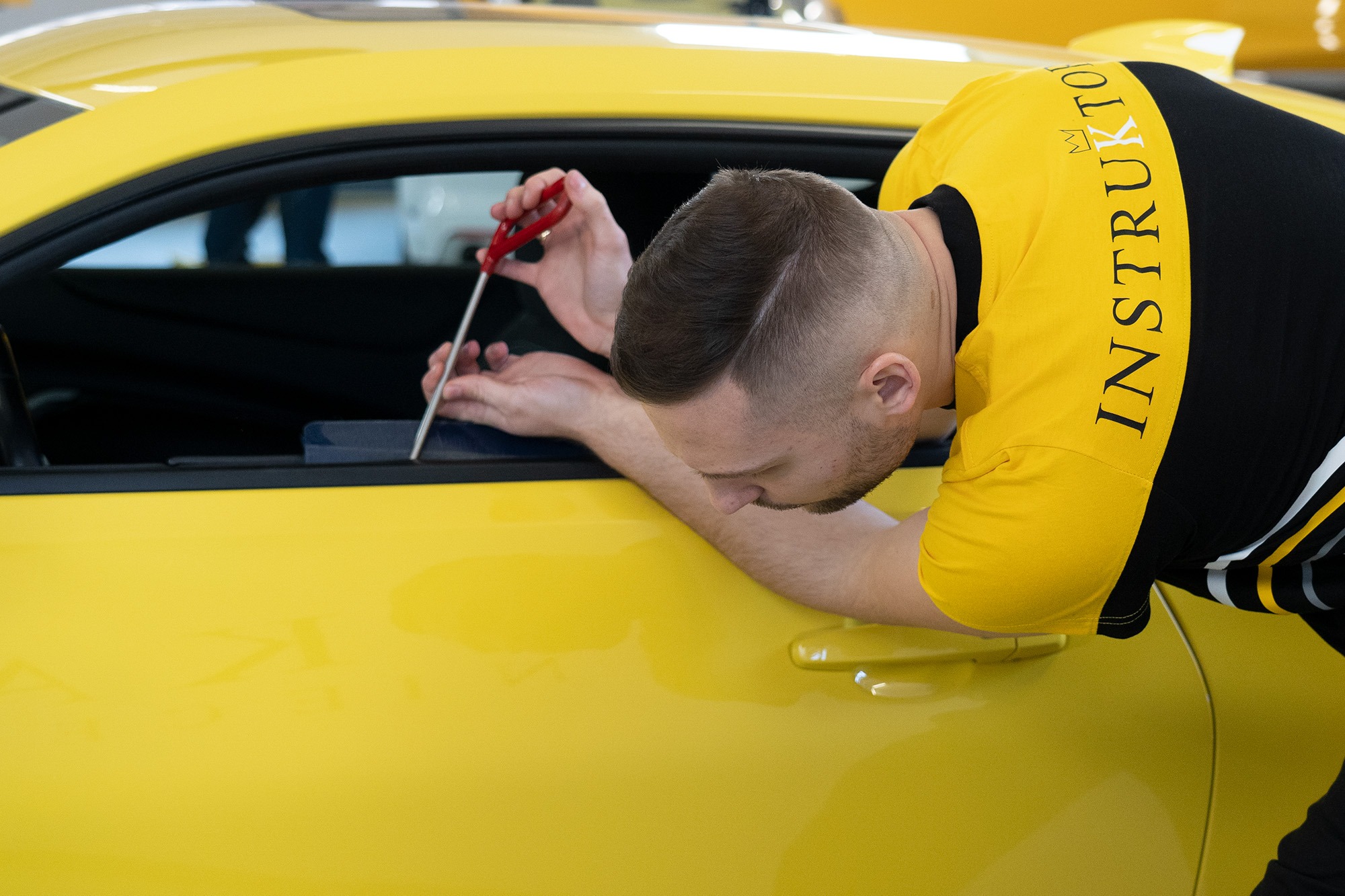Mężczyzna w żółto-czarnej koszulce z napisem 'INSTRUKTOR' delikatnie podważa uszczelkę okna w żółtym samochodzie za pomocą cienkiego narzędzia z czerwoną rączką.