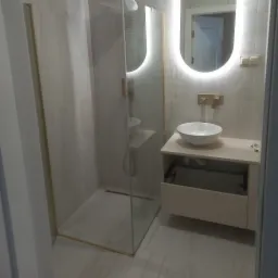 Nowoczesna łazienka z kabiną prysznicową ze złotymi akcentami, owalnym lustrem z podświetleniem LED i białą umywalką nablatową na beżowej szafce z otwartą szufladą.