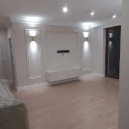 Jasne, puste wnętrze po remoncie, z panelami podłogowymi, dekoracyjnymi listwami ściennymi, oświetleniem punktowym i meblami RTV pod telewizorem.