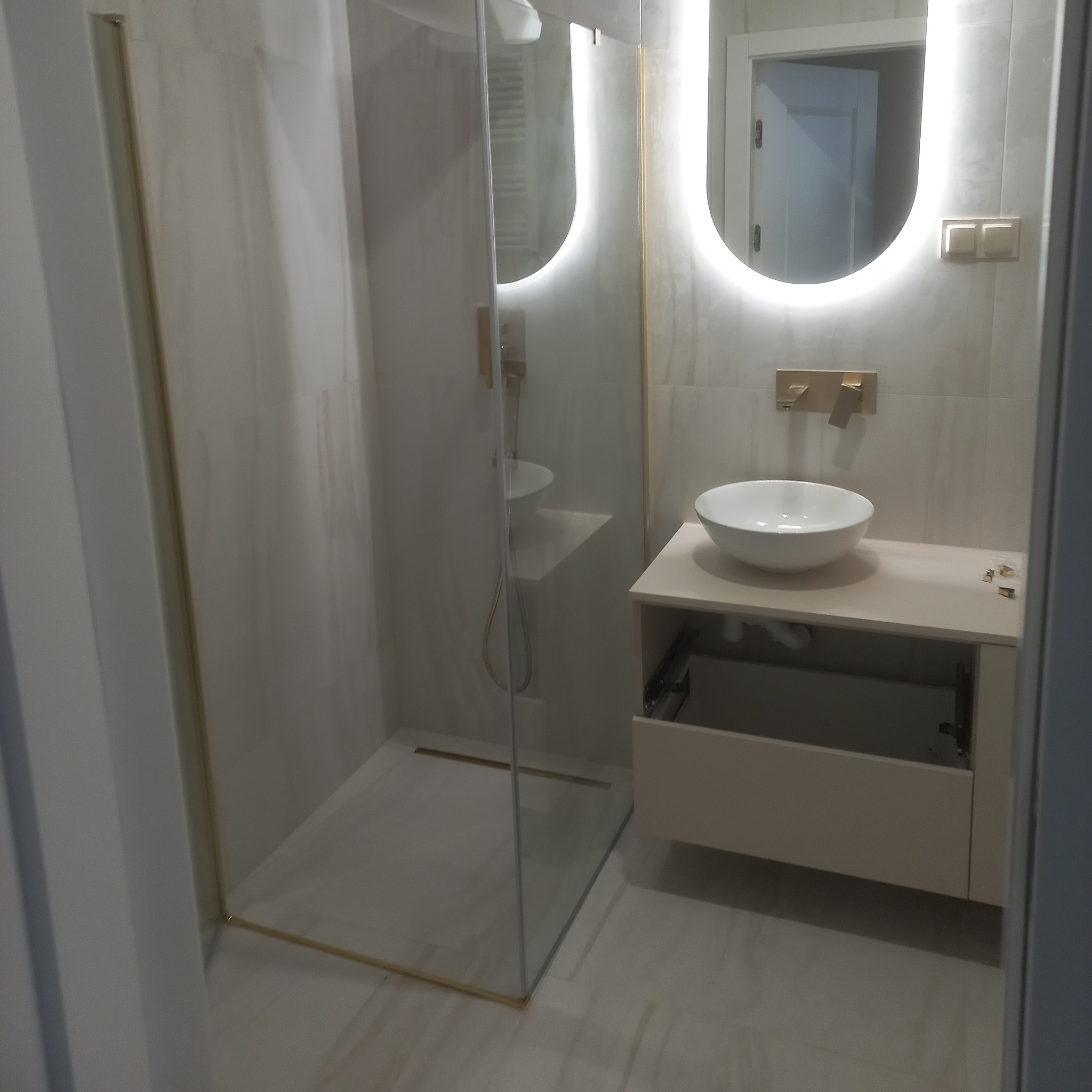 Nowoczesna łazienka z kabiną prysznicową ze złotymi akcentami, owalnym lustrem z podświetleniem LED i białą umywalką nablatową na beżowej szafce z otwartą szufladą.