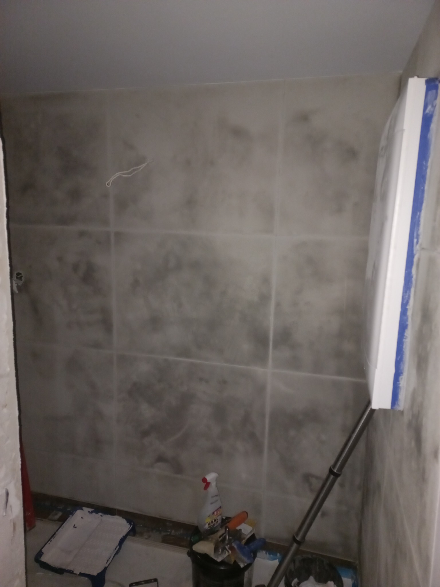 Ściana pokryta szarym, wzorzystym tynkiem dekoracyjnym, widoczny wałek malarski z niebieską taśmą zabezpieczającą, wiaderko z narzędziami i kuweta z farbą na podłodze.