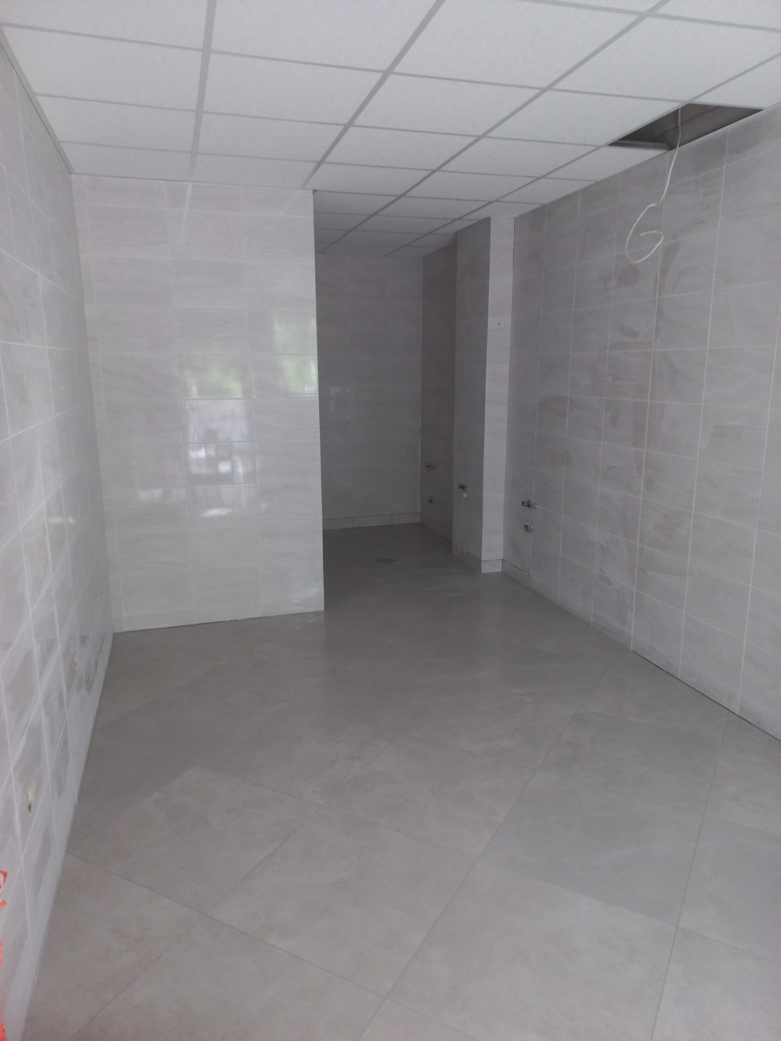 Puste, wykafelkowane pomieszczenie z otwartym sufitem podwieszanym, ukazujące instalacje. Świeżo położone płytki podłogowe i ścienne w odcieniach szarości.