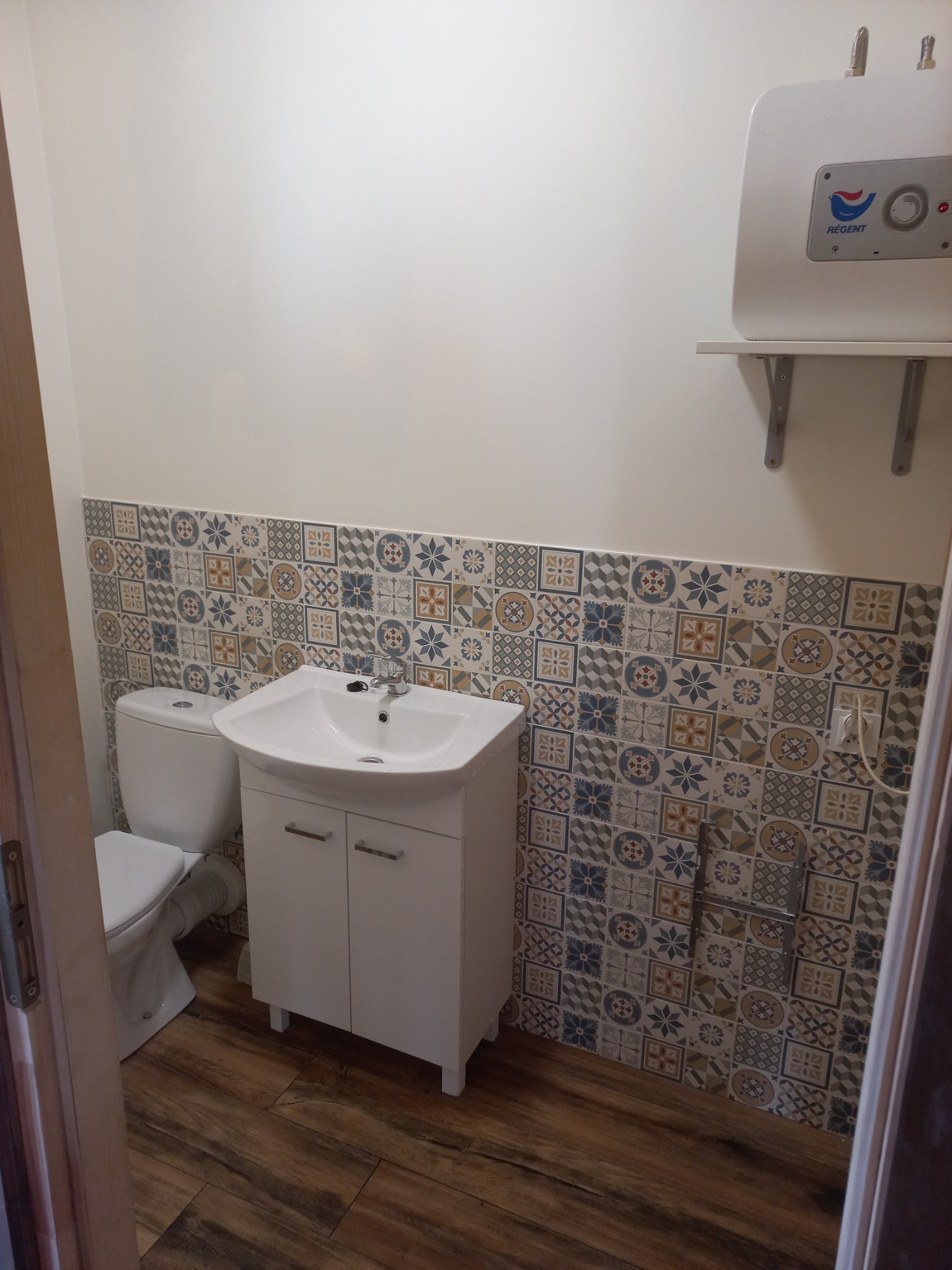Wykończona łazienka z umywalką na szafce, toaletą i bojlerem na ścianie z dekoracyjnymi kaflami, widoczne drewnopodobne panele podłogowe i fragment ościeżnicy.