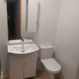 Usługi remontowe Marcin Wyrzykowski - Wykończona łazienka z białą armaturą: umywalka z szafką, toaleta i lustro z szafką wiszącą na białych płytkach, widoczne drewnopodobne panele podłogowe.