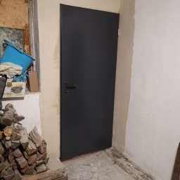 Usługi remontowe Marcin Wyrzykowski - Nowo zamontowane, grafitowe drzwi wewnętrzne z czarną klamką w surowym pomieszczeniu podczas remontu, widoczne ślady po gładzi na ścianach i sterta drewna obok.