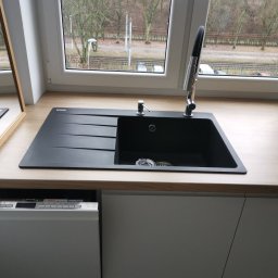 Usługi remontowe Marcin Wyrzykowski - Czarny, prostokątny zlew kuchenny z ociekaczem i chromowaną baterią, zamontowany w blacie o jasnym, drewnopodobnym wykończeniu, z widoczną częścią zmywarki Bosch pod spodem i oknem w tle.