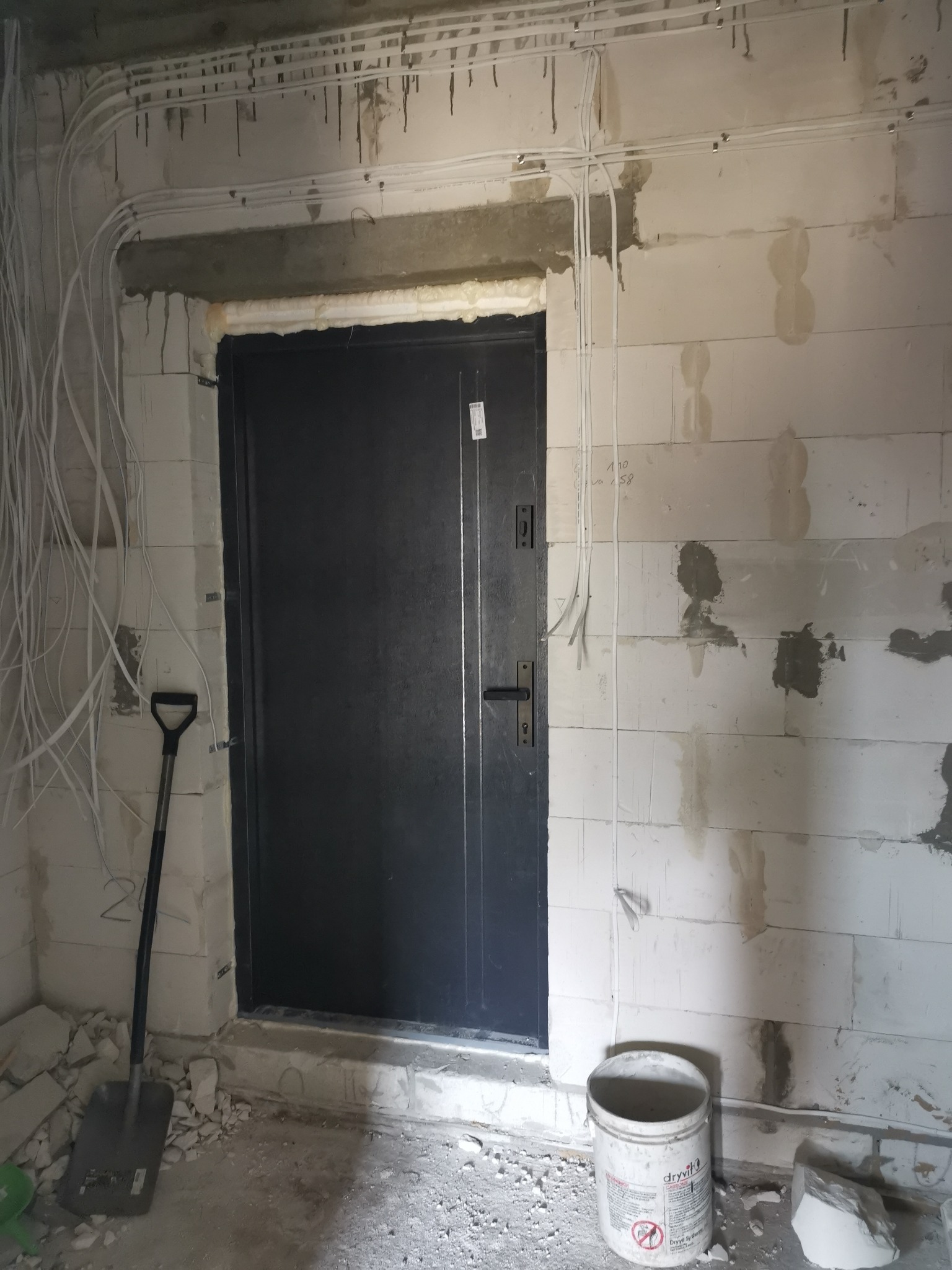 Ciemne drzwi wejściowe zamontowane w surowym stanie, widoczne przewody elektryczne, ślady po zaprawie na ścianach z bloczków, wiadro po kleju i łopata.