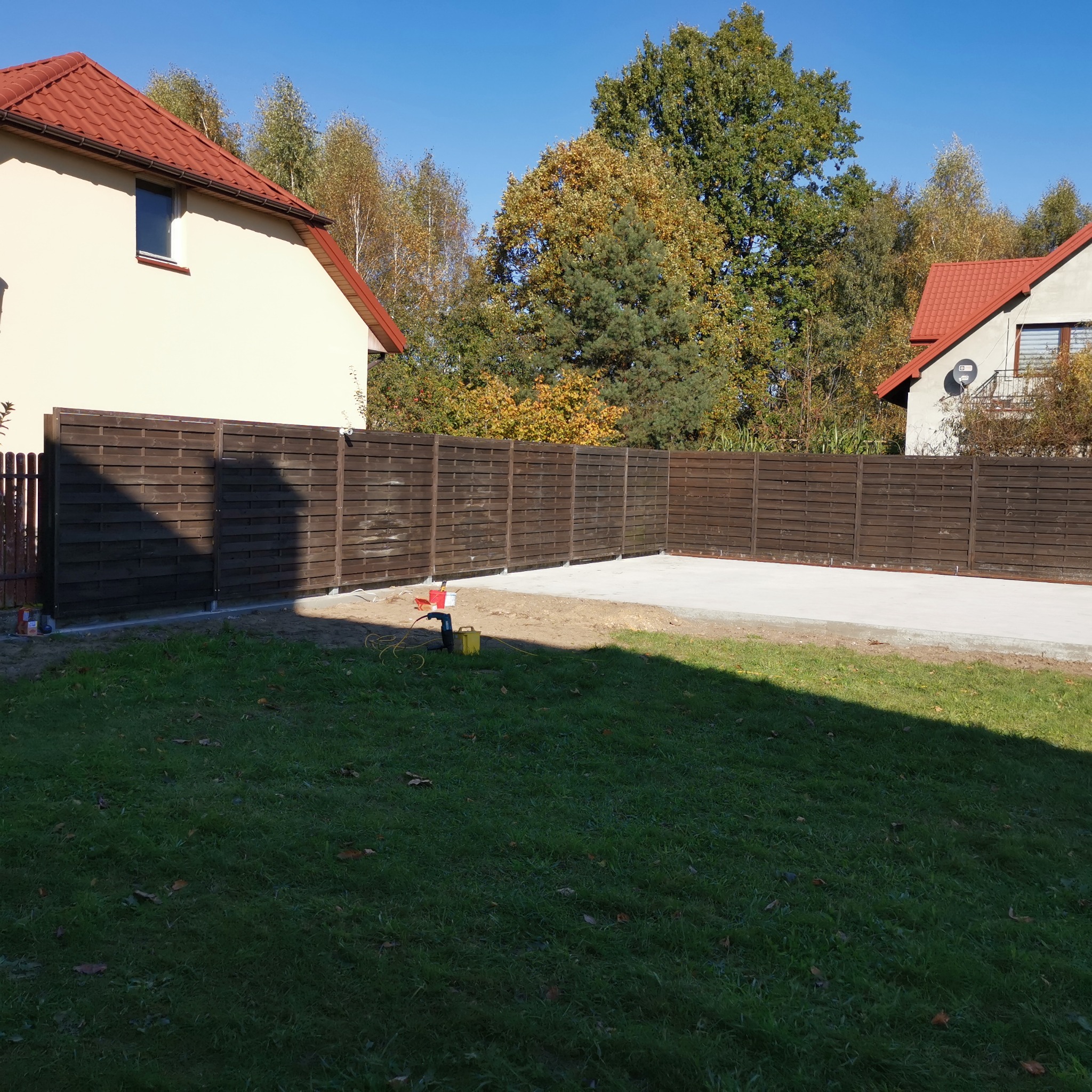 Nowo postawione, ciemnobrązowe, drewniane ogrodzenie panelowe na betonowej podmurówce, widok z trawnika w słoneczny dzień, z narzędziami budowlanymi w tle.