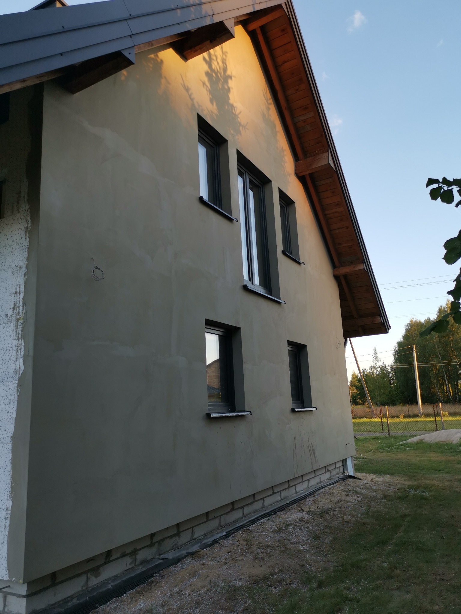 Świeżo otynkowana ściana domu jednorodzinnego z zamontowanymi oknami, widoczne niedoskonałości w fakturze tynku, fragment ocieplenia na rogu budynku i podmurówka z cegły.