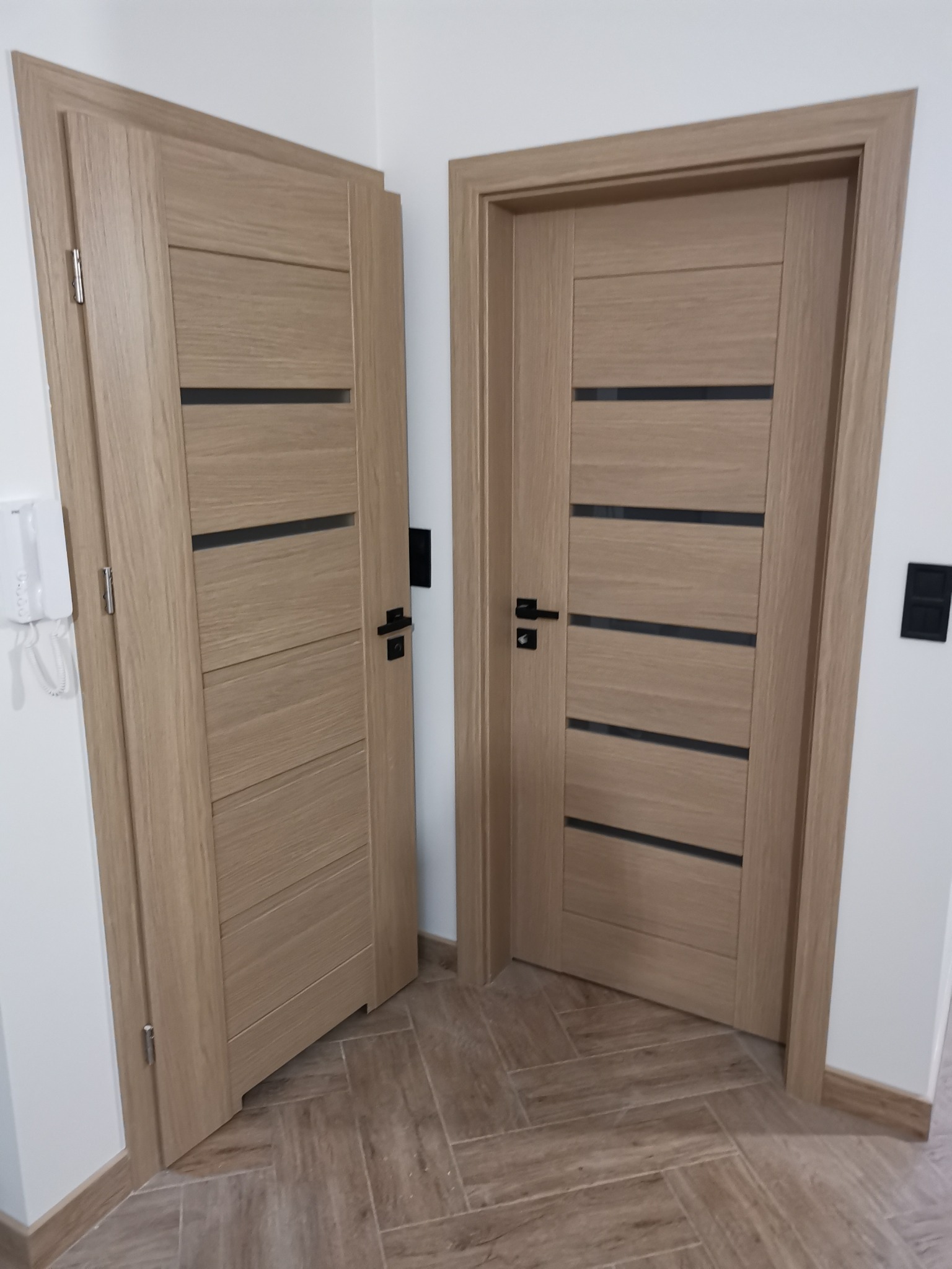 Dwa nowoczesne, drewniane drzwi wewnętrzne z czarnymi, poziomymi wstawkami, zamontowane w jasnym pomieszczeniu z podłogą wyłożoną płytkami imitującymi drewno ułożonymi w jodełkę.