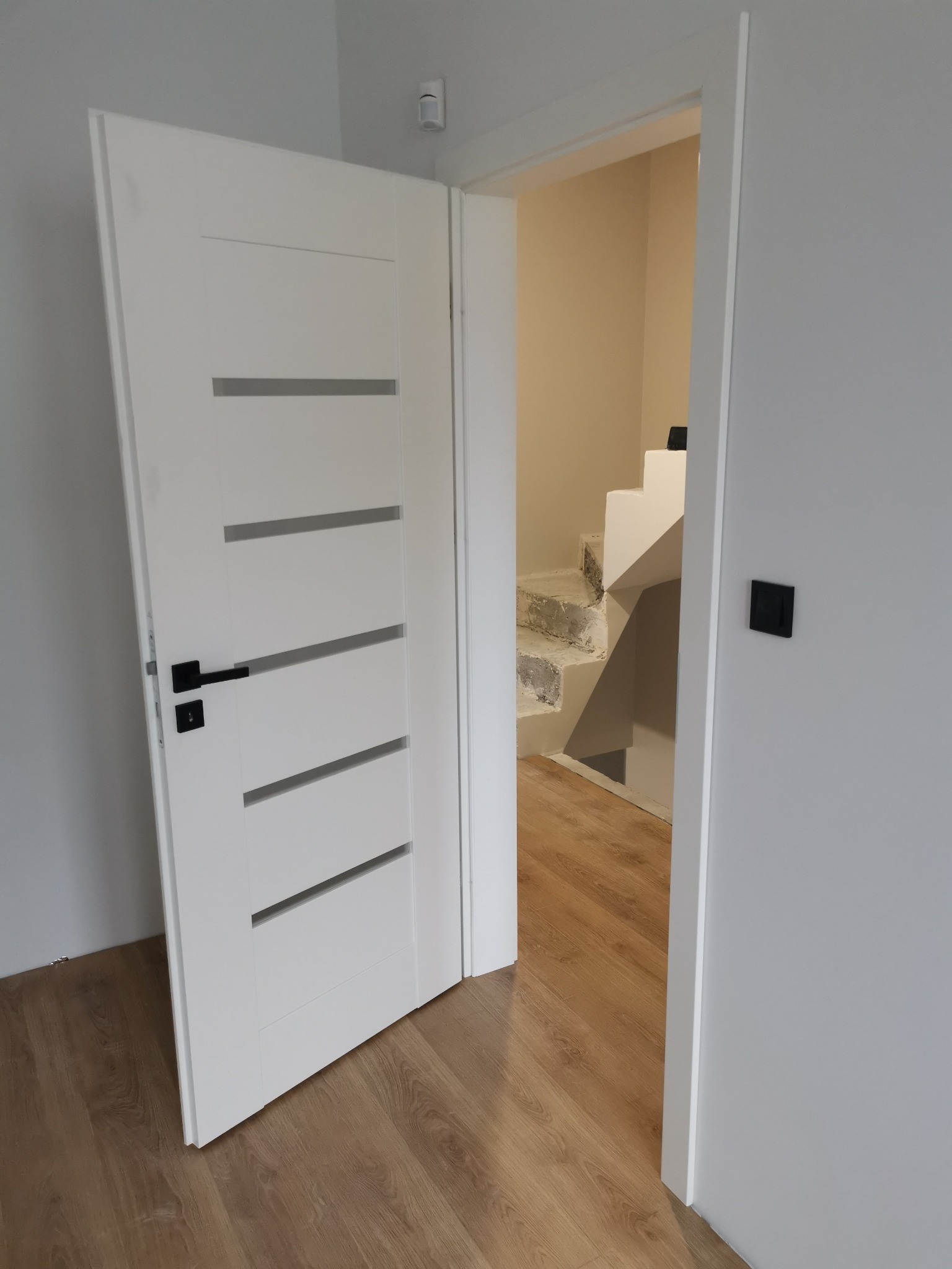 Otwarta biała, nowoczesna wewnętrzna drzwi z poziomymi przeszkleniami i czarną klamką, widok na schody w trakcie remontu, podłoga drewnopodobna.