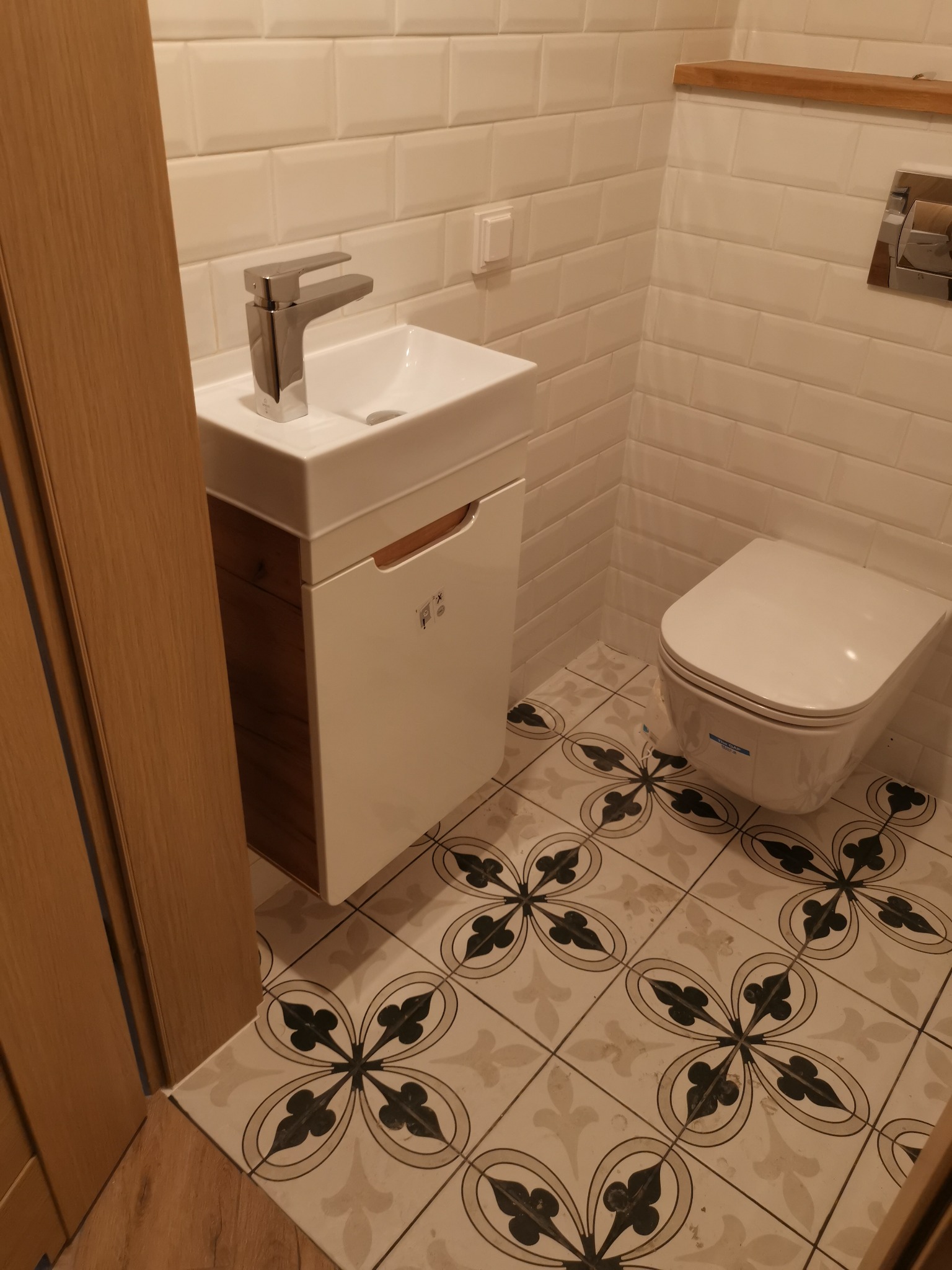 Nowoczesna łazienka z białą armaturą, umywalką z szafką w kolorze drewna, toaletą podwieszaną i płytkami podłogowymi w kwiatowy wzór.