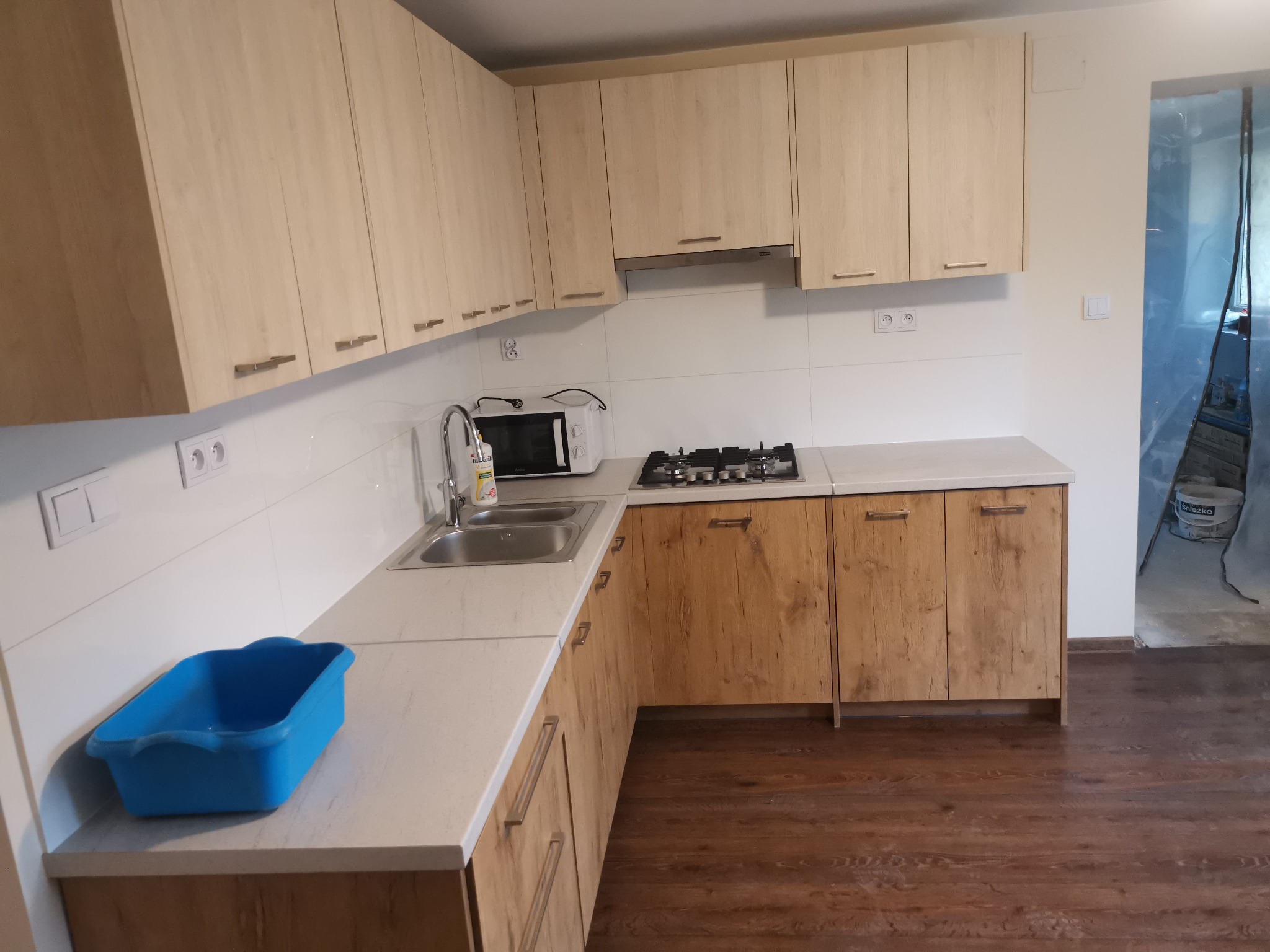Jasna kuchnia z drewnianymi szafkami i białym blatem; w zlewozmywaku płyn do naczyń, a na blacie niebieska miska; widoczny fragment podłogi i zabezpieczone folią malarską przejście do innego...