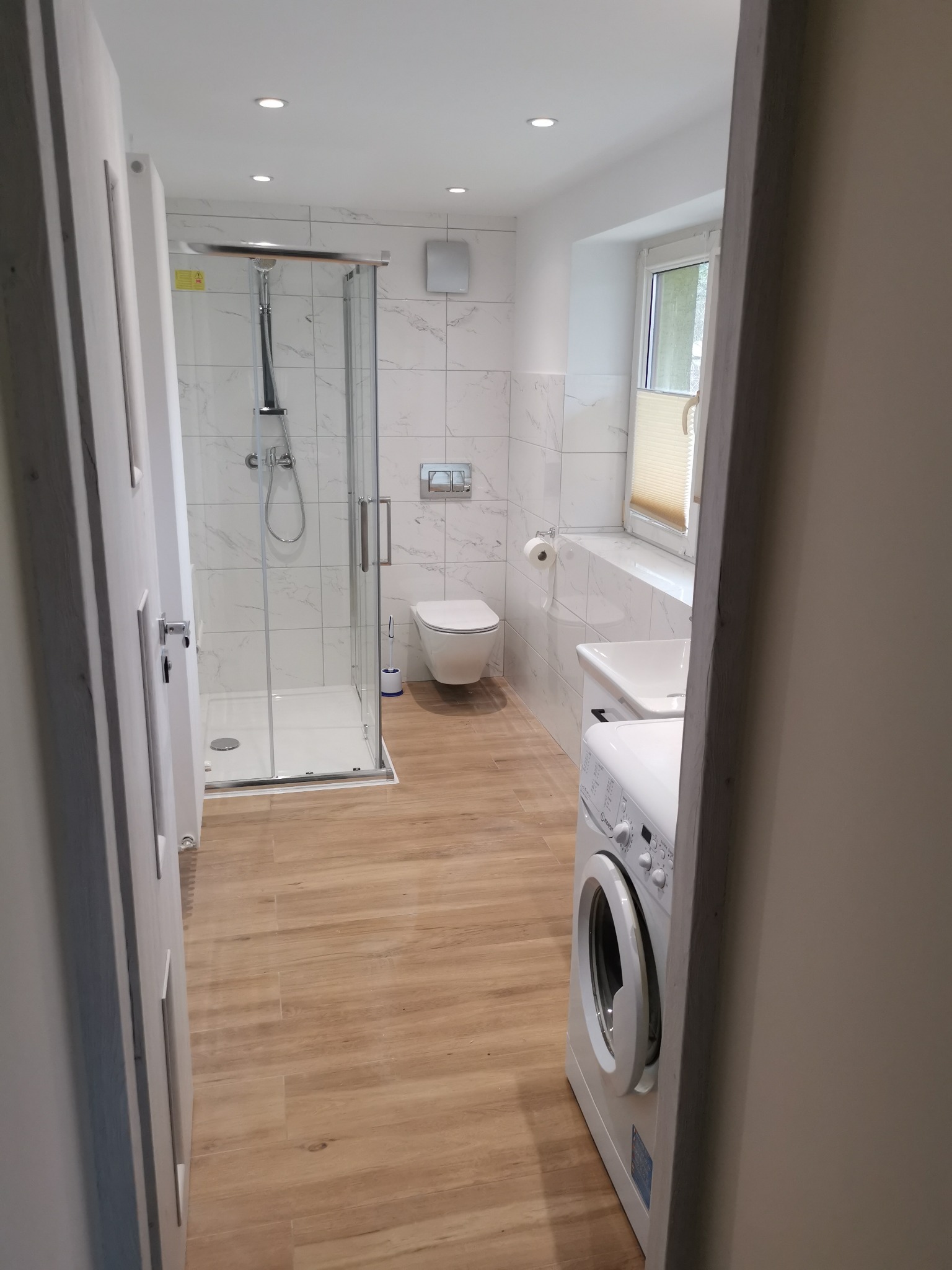 Jasna łazienka z kabiną prysznicową, toaletą, umywalką i pralką, wykończona białymi płytkami imitującymi marmur i drewnopodobną podłogą, widziana zza uchylonych drzwi.
