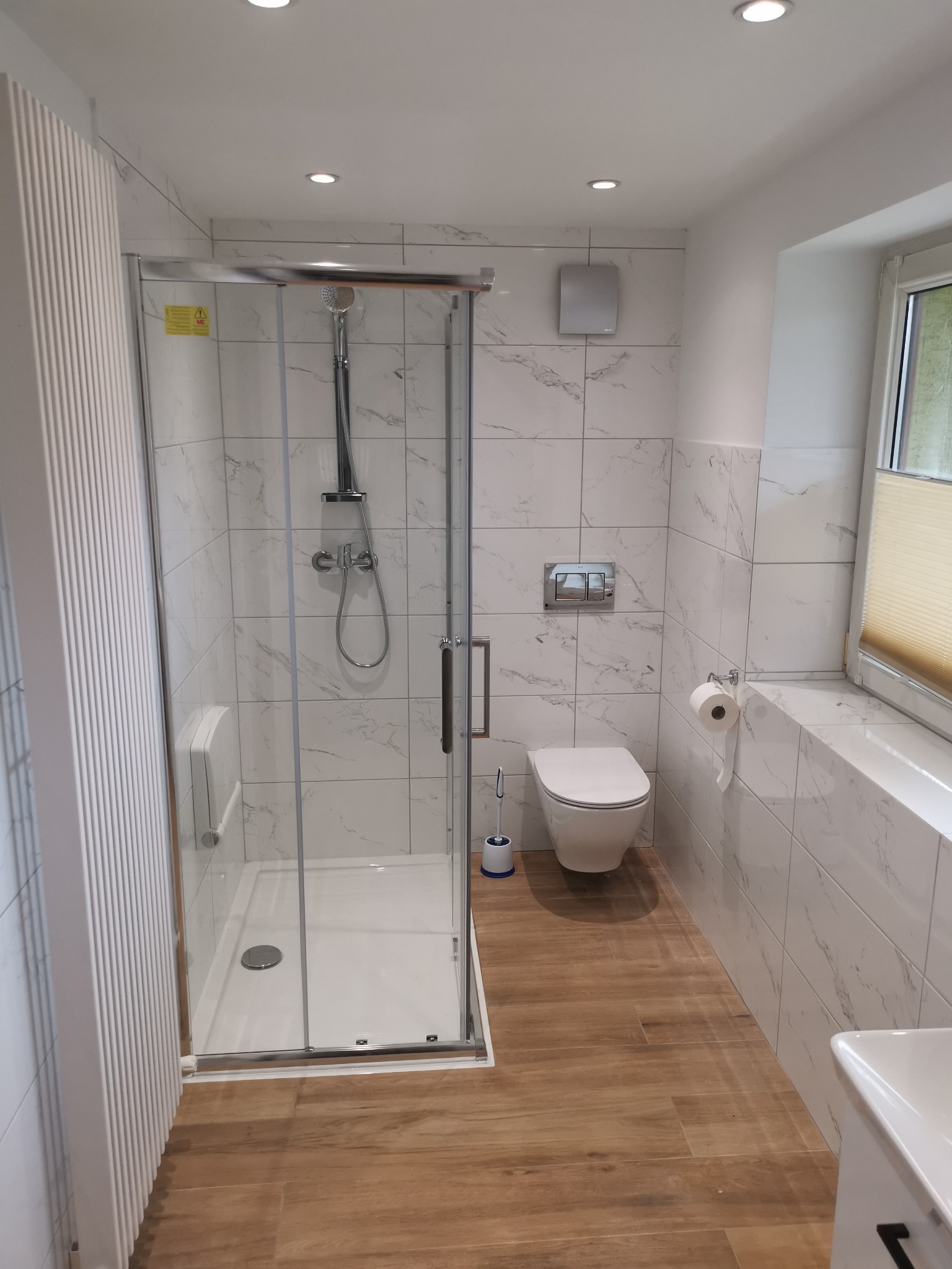 Nowoczesna łazienka z kabiną prysznicową ze szklanymi drzwiami, wiszącą toaletą, grzejnikiem drabinkowym, marmurowymi płytkami na ścianach i panelami podłogowymi imitującymi drewno.