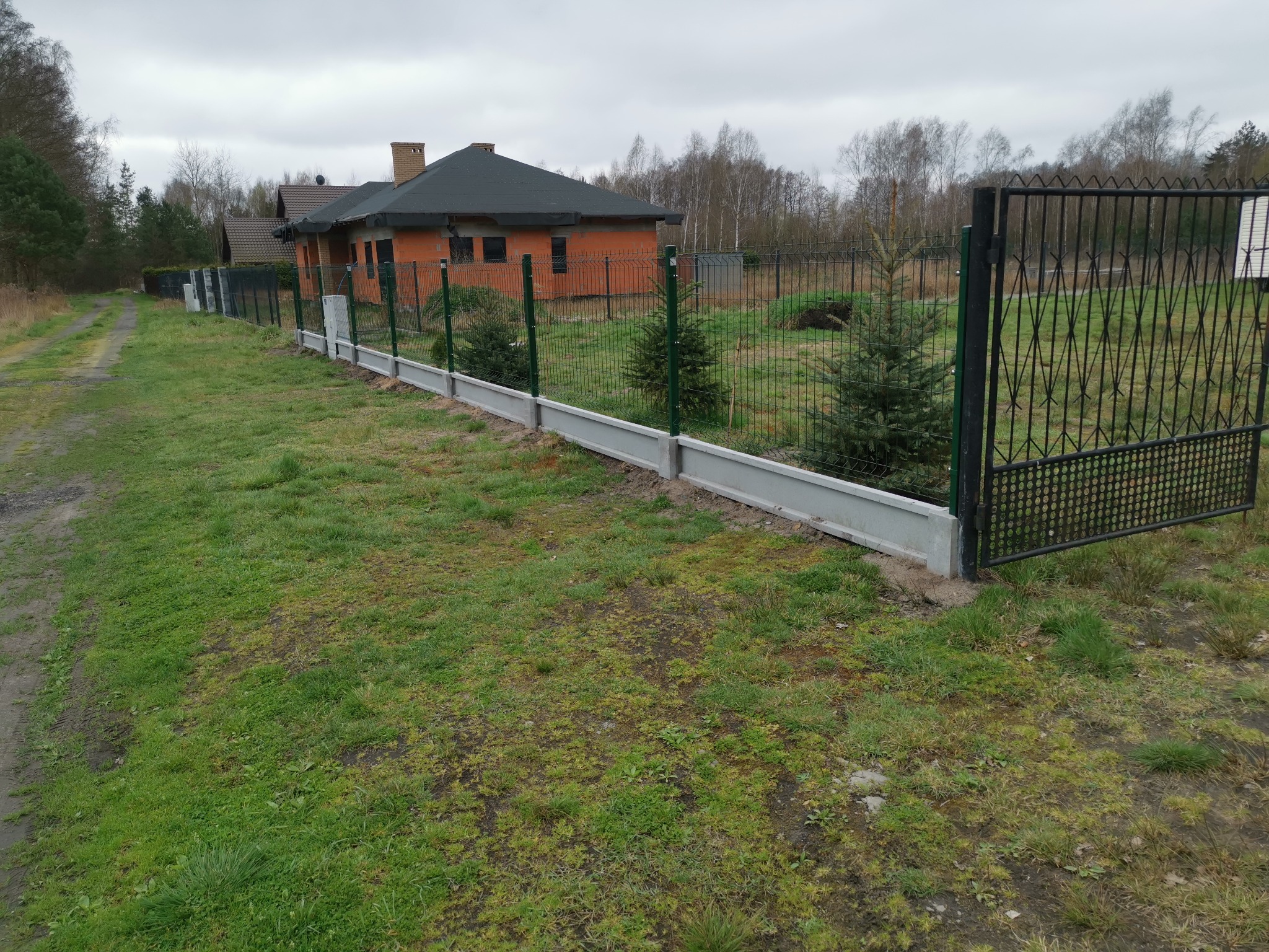 Nowoczesne ogrodzenie panelowe z podmurówką betonową i metalową bramą wjazdową, na tle domu w trakcie budowy i zielonego trawnika.