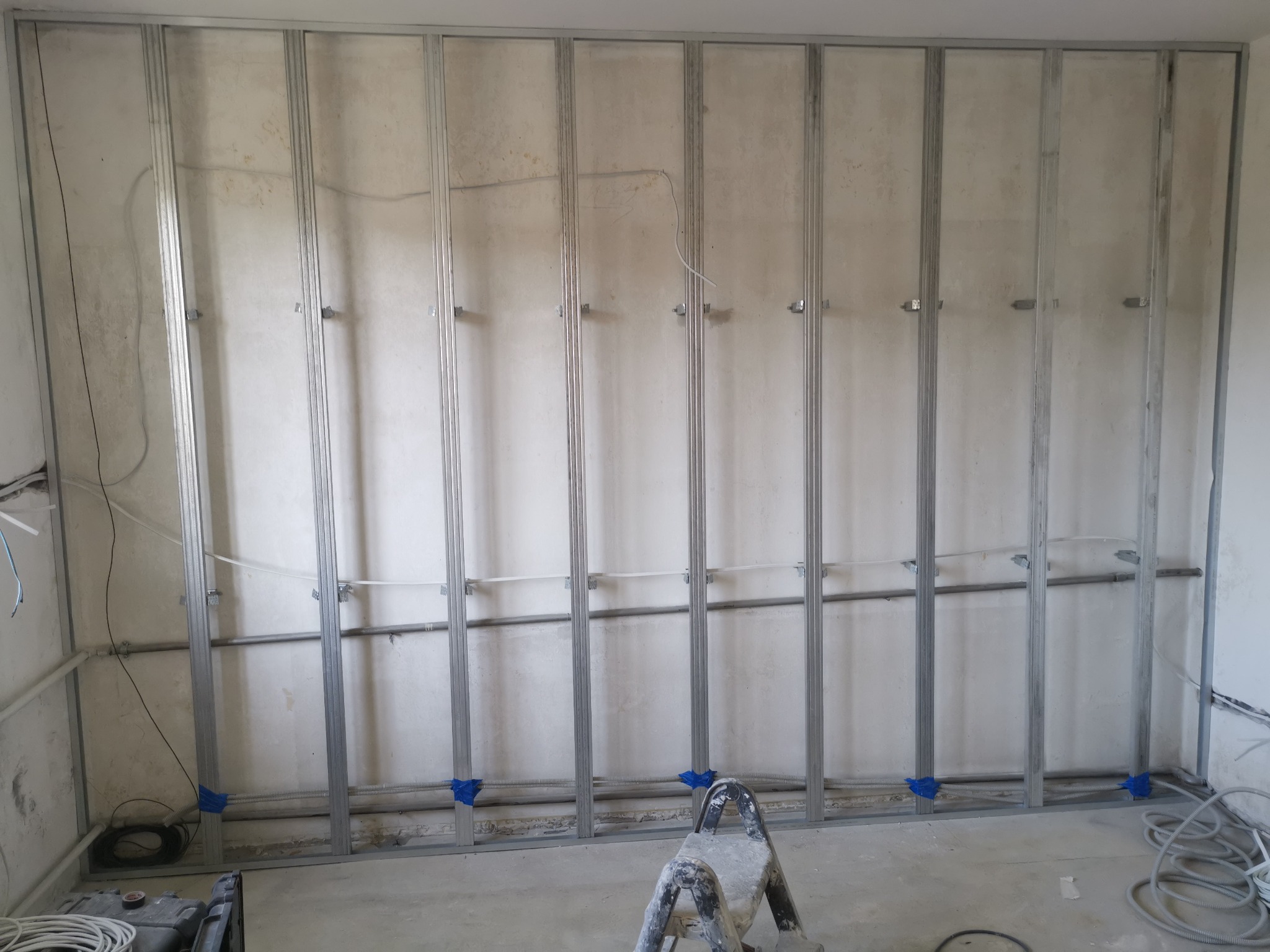 Stelaż metalowy pod zabudowę ścian gipsowo-kartonowych z widocznymi przewodami elektrycznymi, zabezpieczonymi niebieską taśmą, oraz drabina malarska w pomieszczeniu w trakcie remontu.