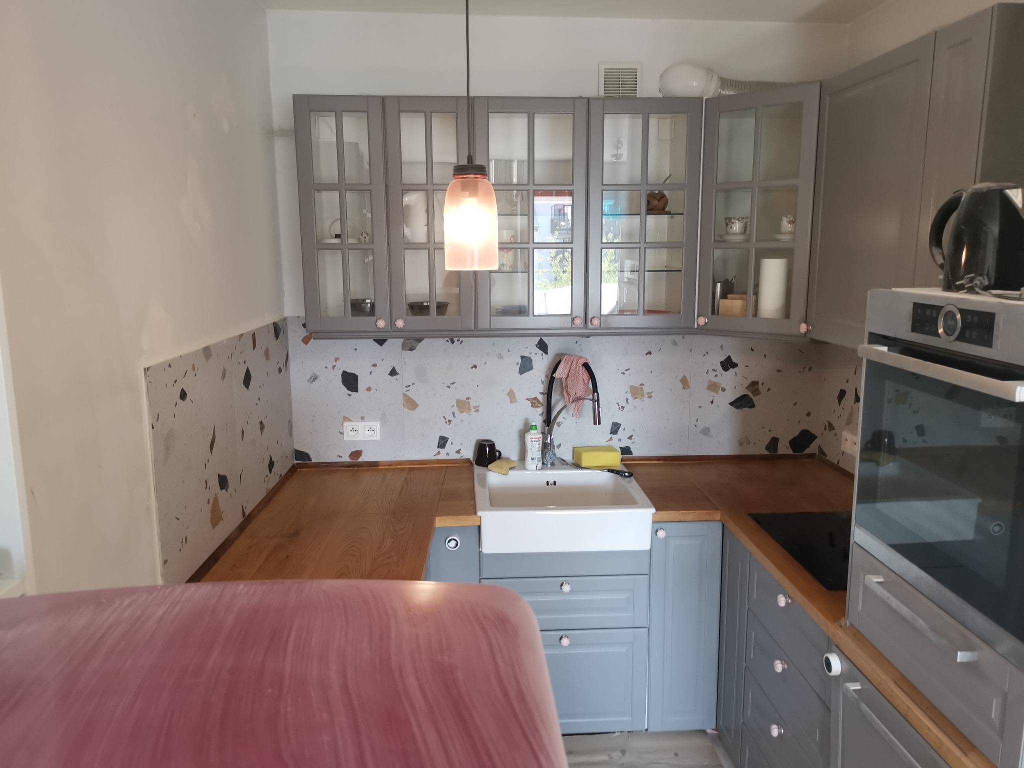 Wykończona kuchnia z szarymi szafkami, drewnianym blatem i mozaikową ścianą nad zlewem. Nad zlewem wisi lampa w kształcie słoika, a na blacie stoją płyny i gąbka do mycia naczyń.