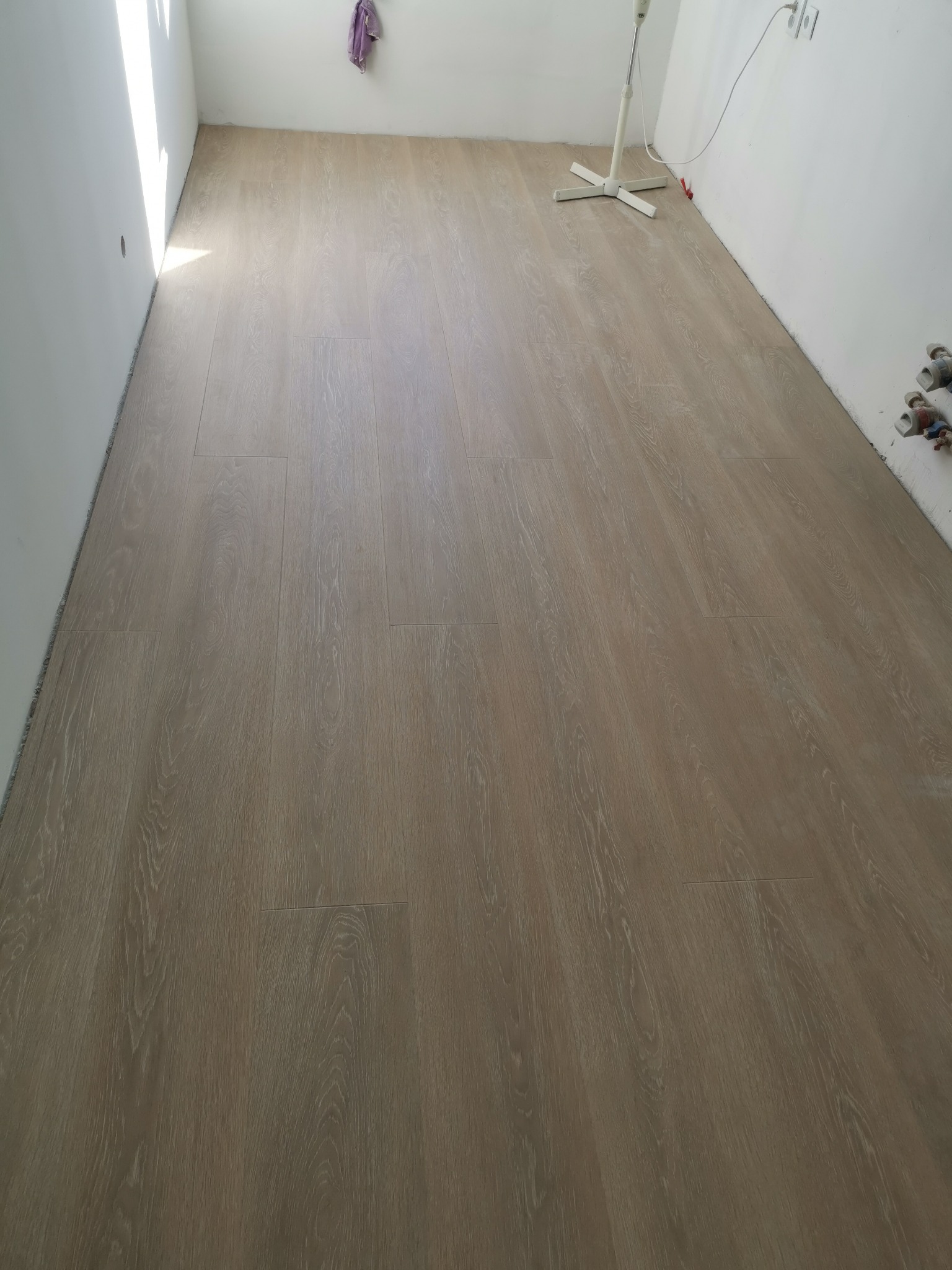 Ułożone panele podłogowe w jasnym, pustym pokoju, widoczne wystające rury i gniazdka elektryczne przy ścianie.