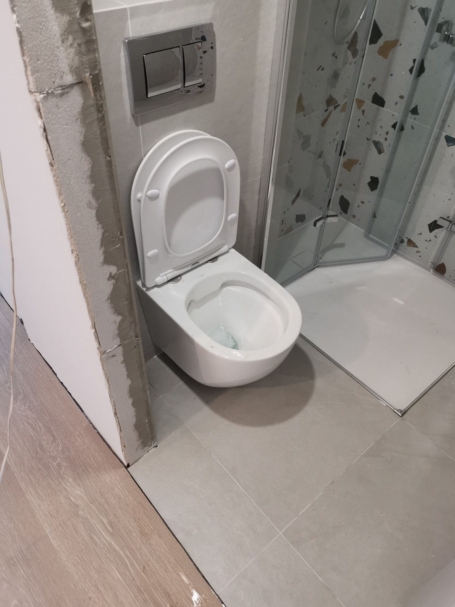 Wisząca toaleta z otwartą klapą, zamontowana w łazience z szarą glazurą i kabiną prysznicową z mozaiką, widoczne niedokończone wykończenie ściany.