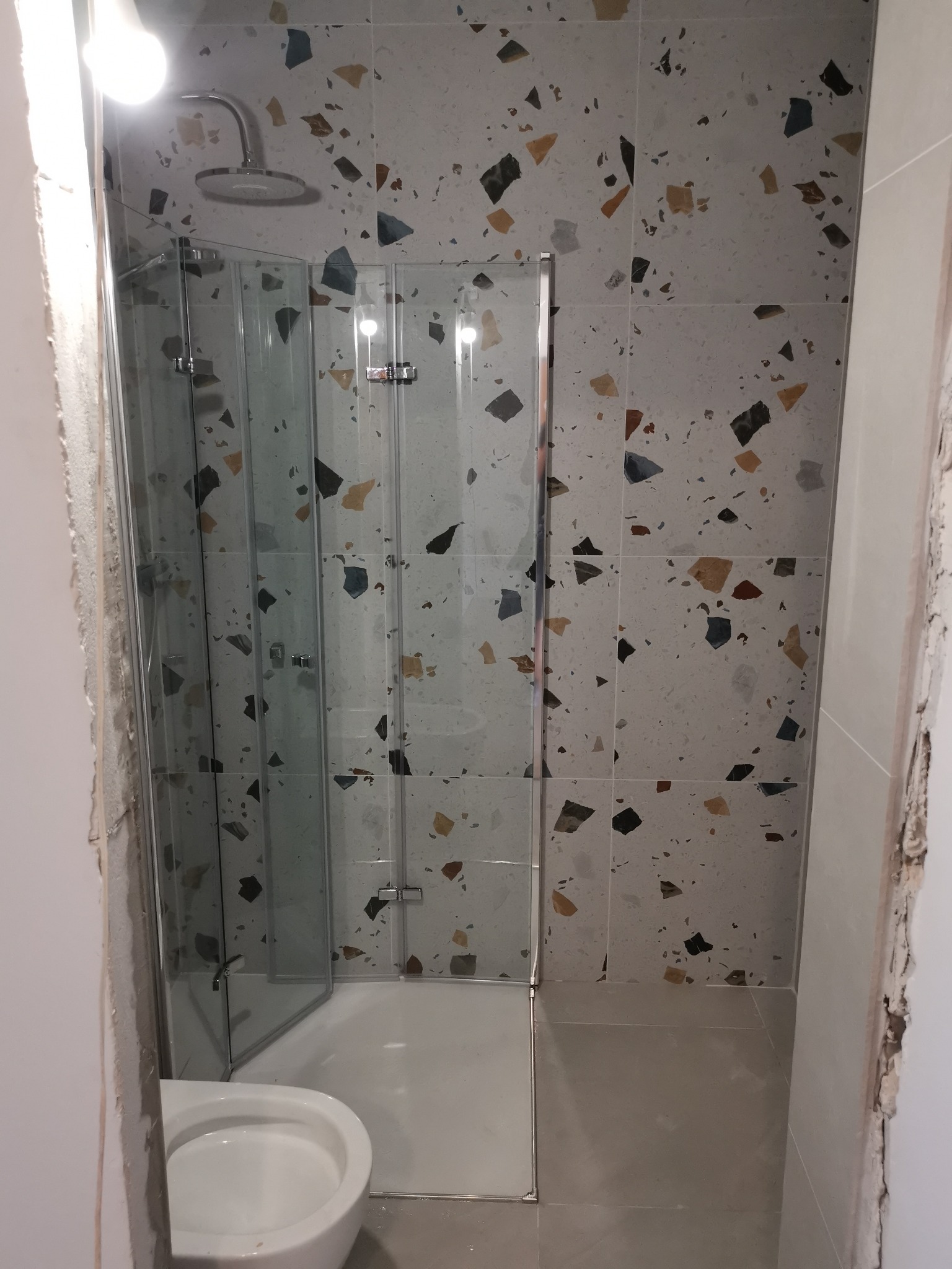 Nowoczesna kabina prysznicowa z rozkładanymi drzwiami, zamontowana w łazience w Tłuszczu, z płytkami imitującymi lastryko na ścianach i widoczną toaletą.