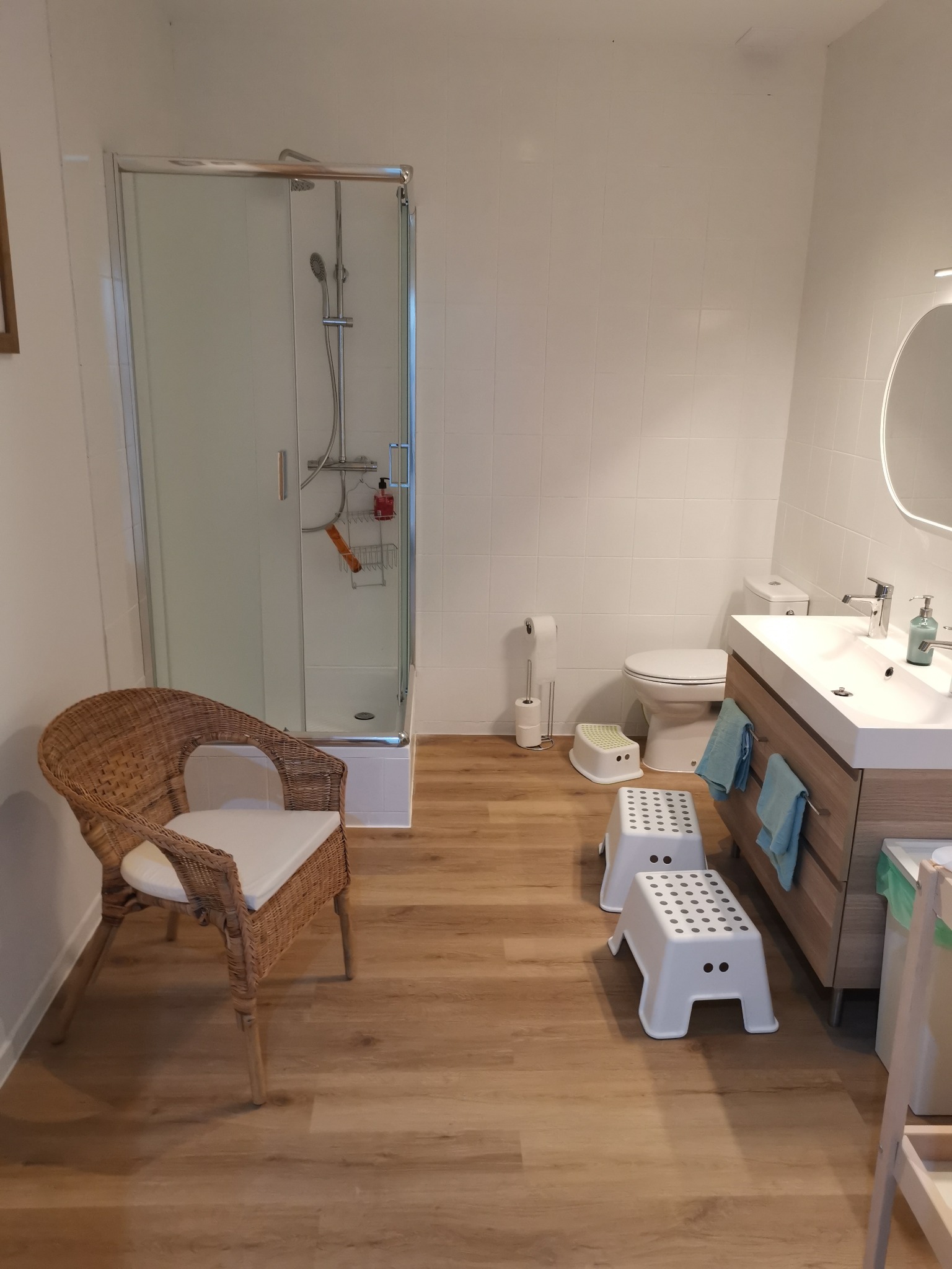 Jasna łazienka z kabiną prysznicową, wiklinowym fotelem z białą poduszką, umywalką z drewnianą szafką, toaletą i dwoma plastikowymi podestami dla dzieci na podłodze imitującej drewno.