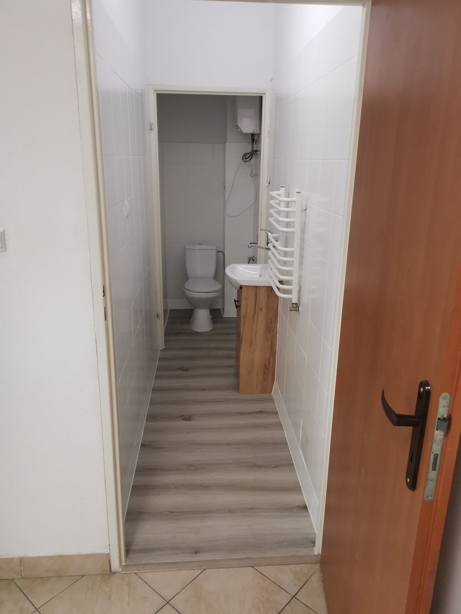 Widok na odnowioną łazienkę z drewnopodobną podłogą, białymi płytkami, toaletą i umywalką z szafką z jasnego drewna, z widocznym grzejnikiem drabinkowym i bojlerem.