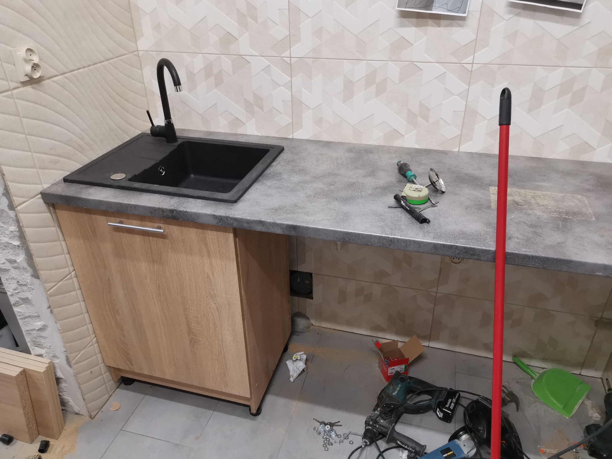 Częściowo zamontowany zlew z czarną baterią na szarym blacie, szafka z jasnego drewna, płytki na ścianie w geometryczny wzór, narzędzia i akcesoria na blacie i podłodze w trakcie remontu łazienki.