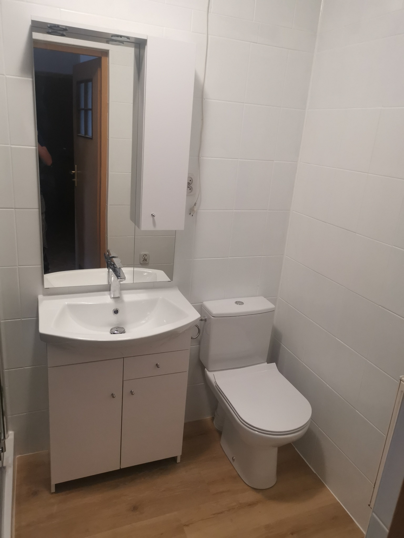 Wykończona łazienka z białą armaturą: umywalka z szafką, toaleta i lustro z szafką wiszącą na białych płytkach, widoczne drewnopodobne panele podłogowe.