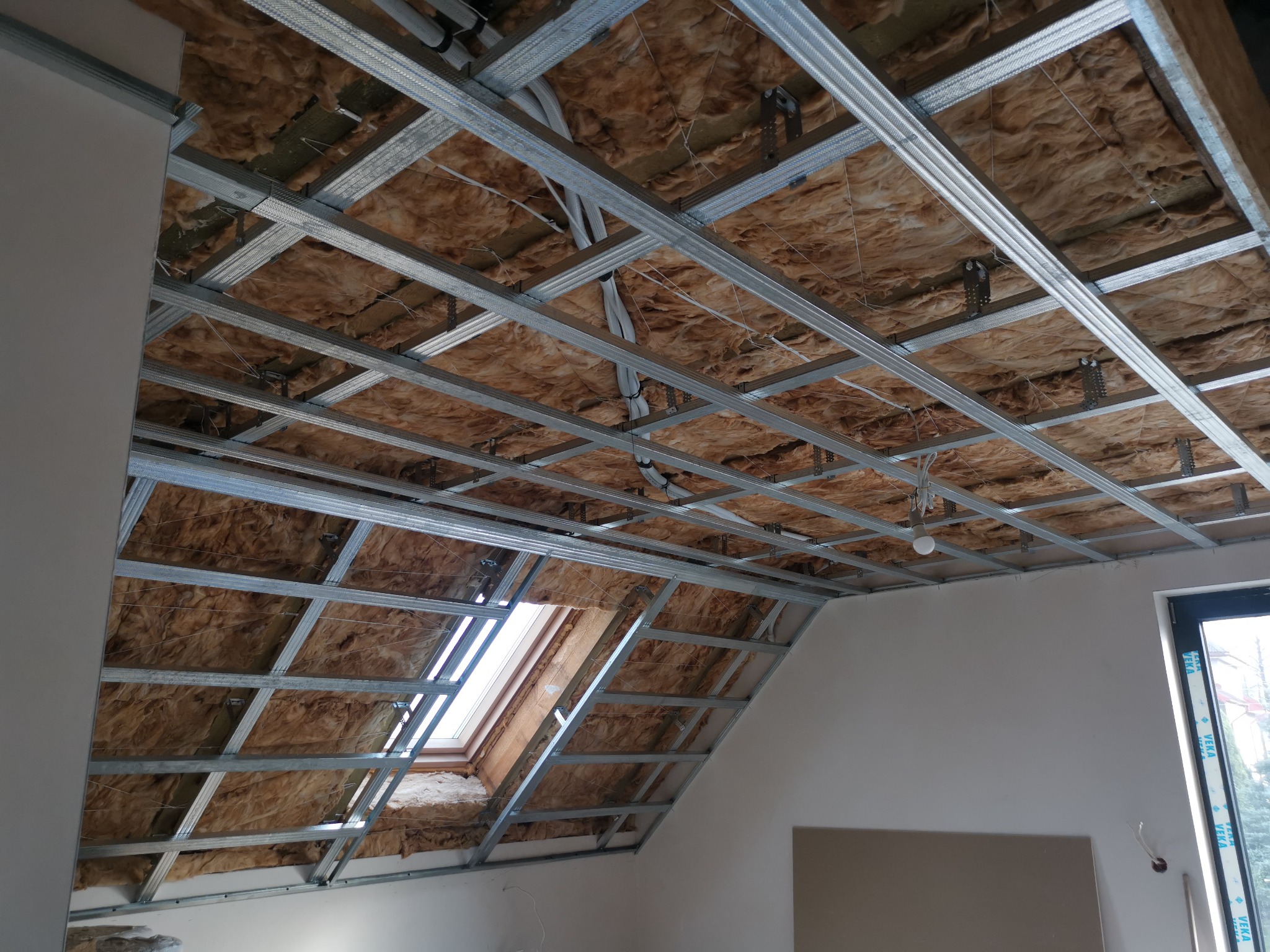 Poddasze w trakcie izolacji: widoczna wełna mineralna między krokwiami i stelaż metalowy pod płyty gipsowo-kartonowe, okno dachowe w skośnej części dachu.