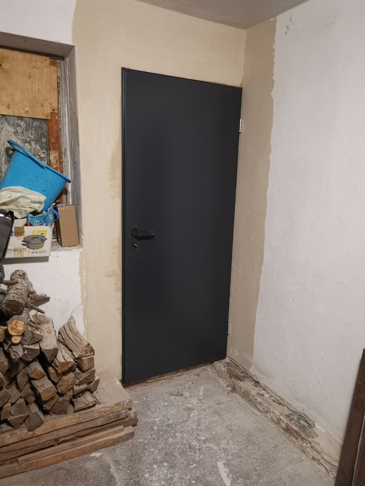 Nowo zamontowane, grafitowe drzwi wewnętrzne z czarną klamką w surowym pomieszczeniu podczas remontu, widoczne ślady po gładzi na ścianach i sterta drewna obok.