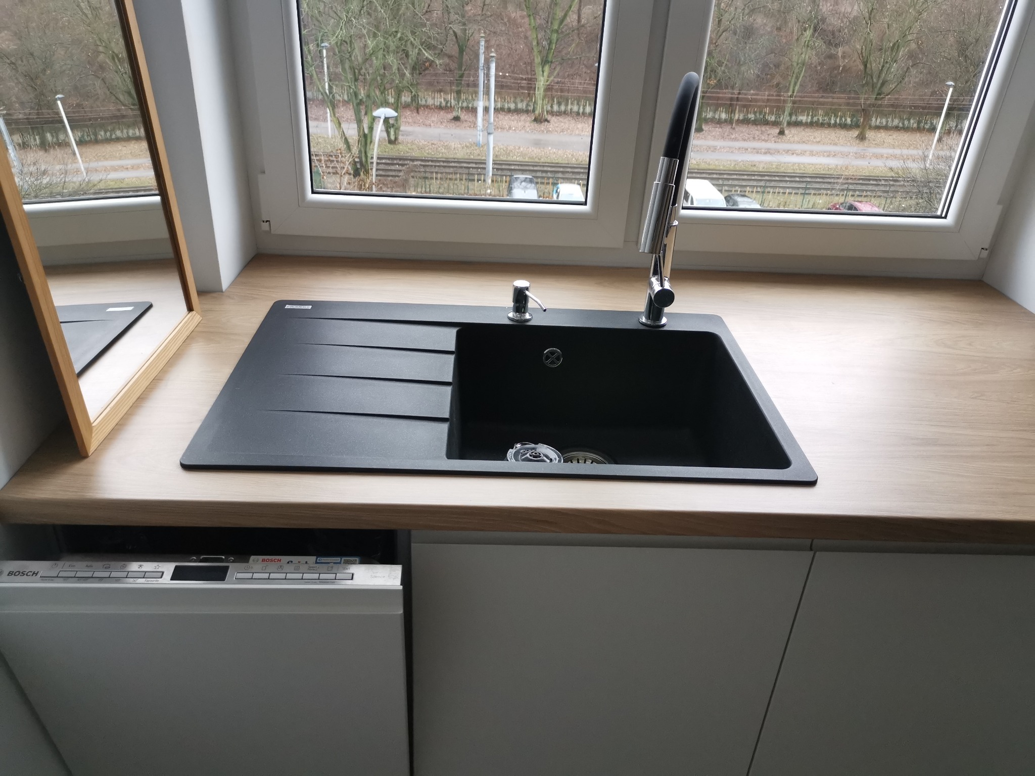 Czarny, prostokątny zlew kuchenny z ociekaczem i chromowaną baterią, zamontowany w blacie o jasnym, drewnopodobnym wykończeniu, z widoczną częścią zmywarki Bosch pod spodem i oknem w tle.