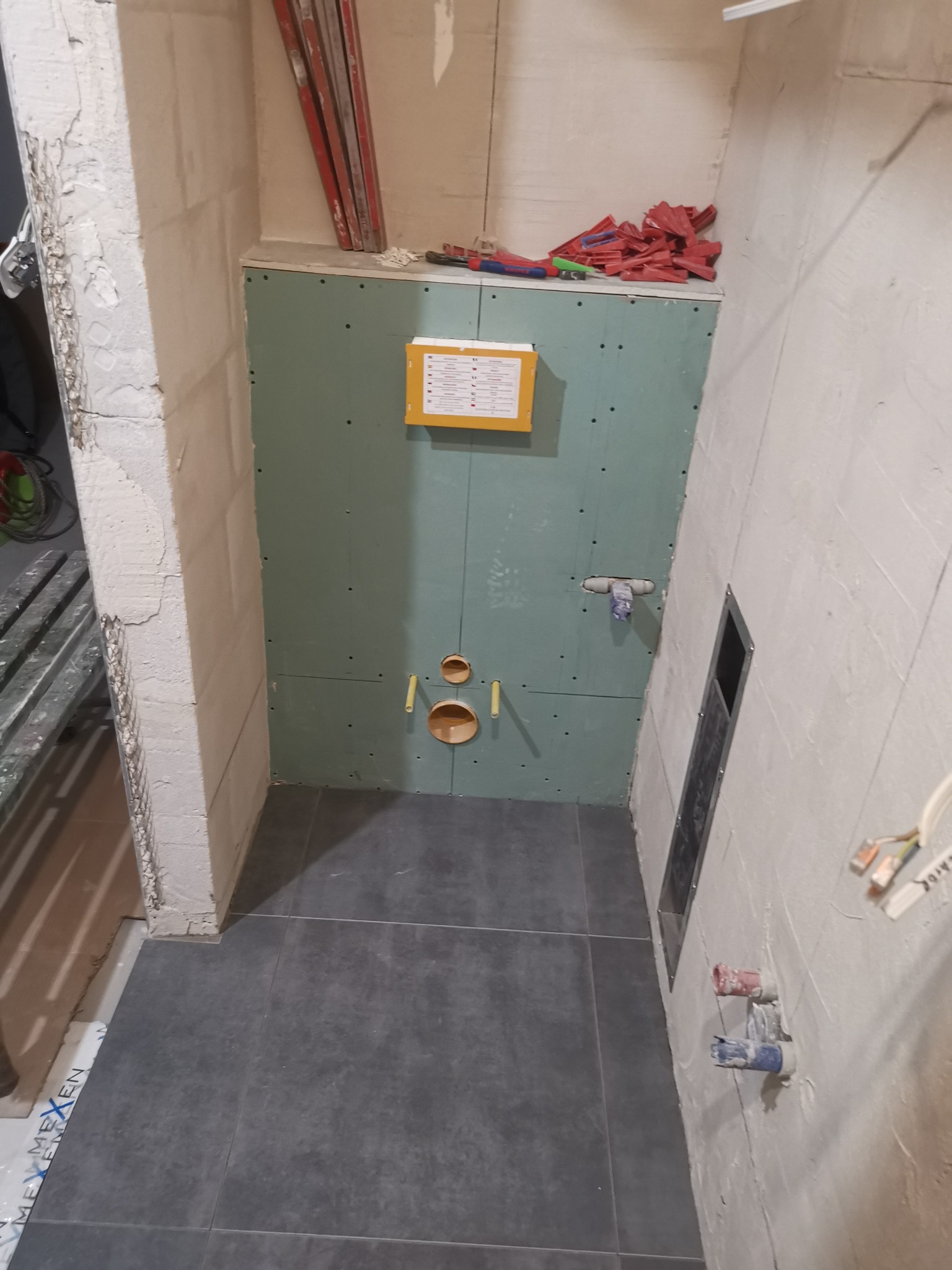 Łazienka w trakcie remontu: widoczne płyty gipsowo-kartonowe zabudowujące stelaż podtynkowy WC, wystające rury instalacji wodnej i kanalizacyjnej, częściowo położone ciemne płytki podłogowe...